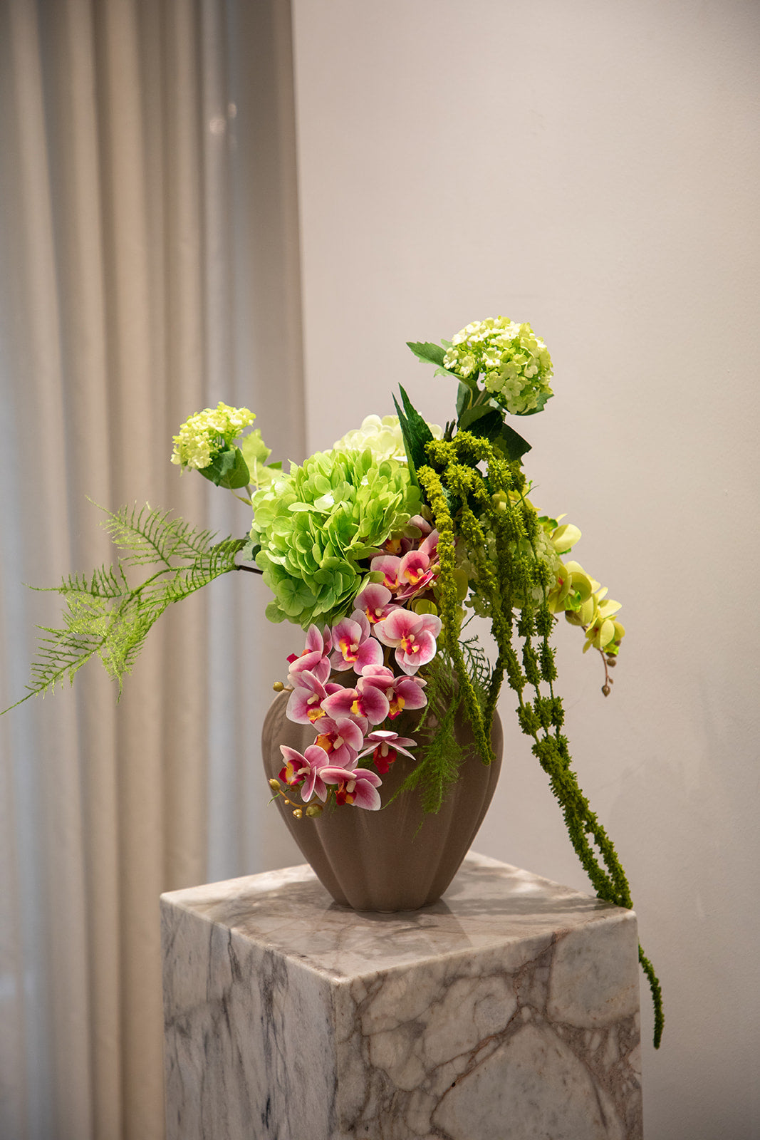 Buket af kunstige blomster i grønne og rosa nuancer. Placeret i en brun keramikvase, der står på en marmor pedestal.