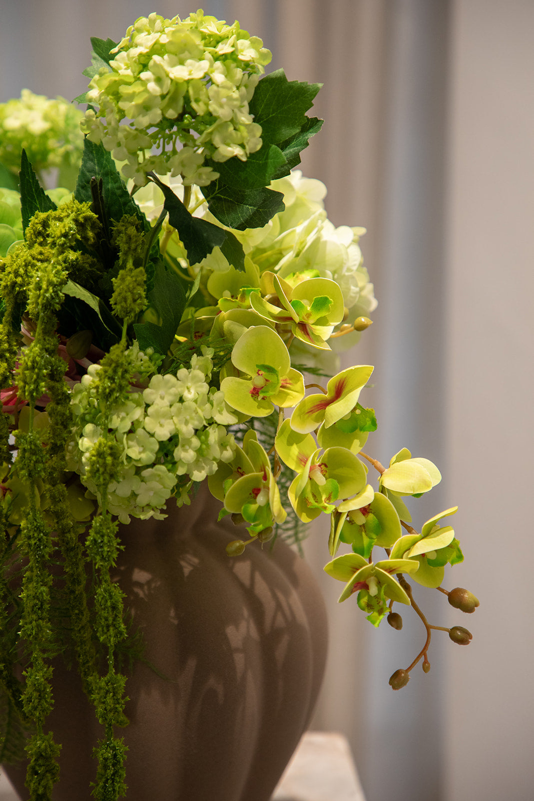 Nærbillede af buket af kunstige blomster i grønne og rosa nuancer. Placeret i en brun keramikvase.