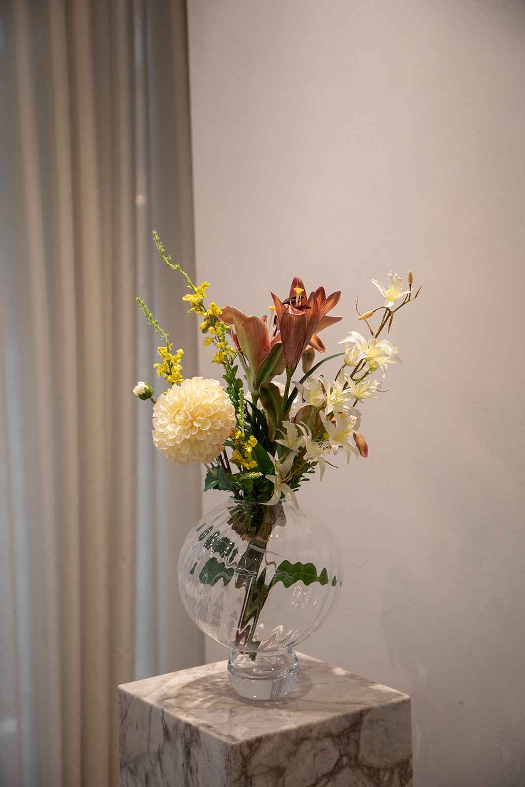 Buket af kunstige blomster i røde, gule og creme farvede nuancer. Placeret i en gennemsigtig glasvase, der står på en marmor pedestal.
