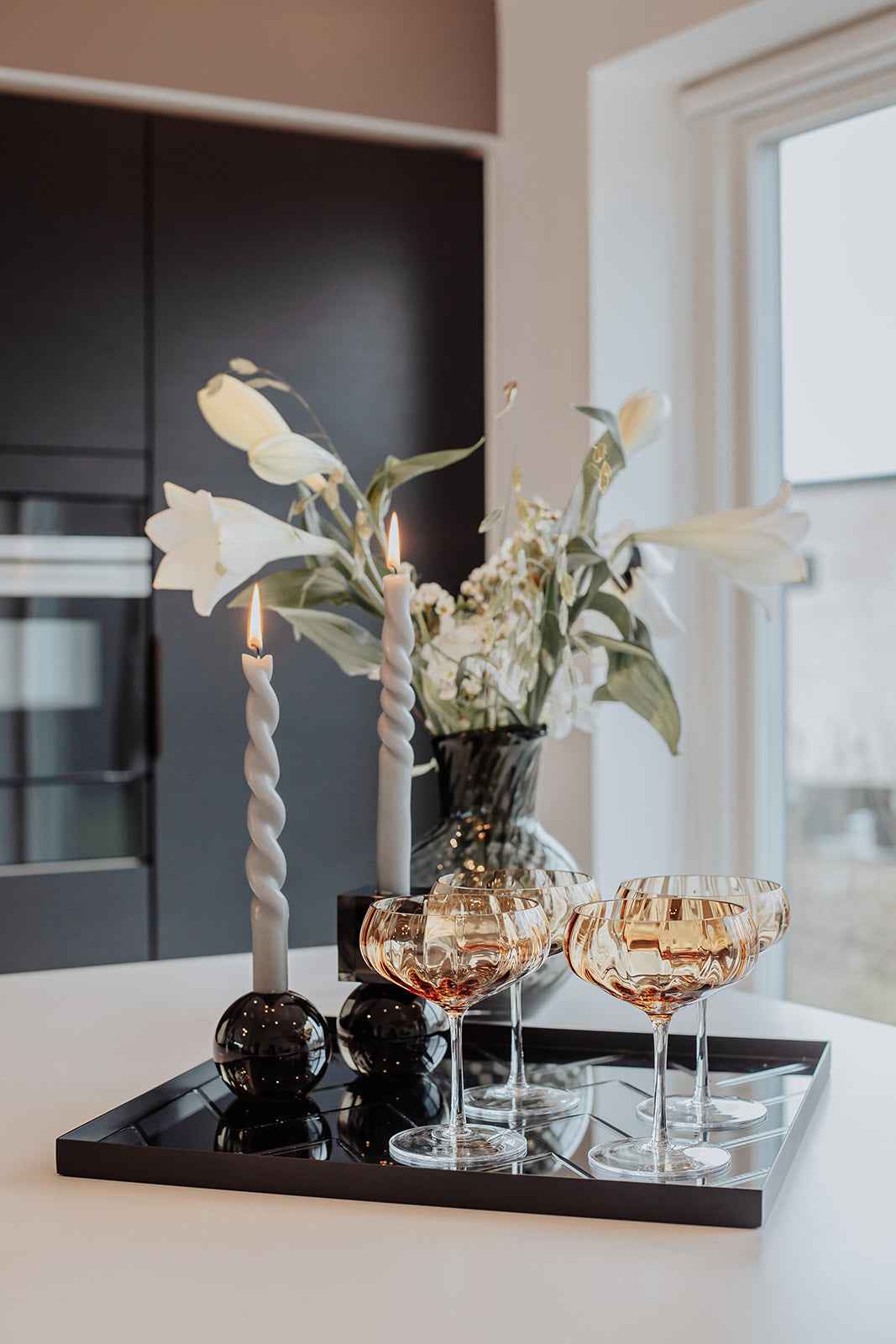 Sort firkantet spejlbakke med diagonalt mønster placeret på en hvid bordplade; på bakken står to tændte snoede stearinlys i mørke glasholdere, tre gyldne champagneglas samt en mørk glasvase med hvide blomster. Baggrunden viser sorte køkkenskabe og et vindue med lysindfald.