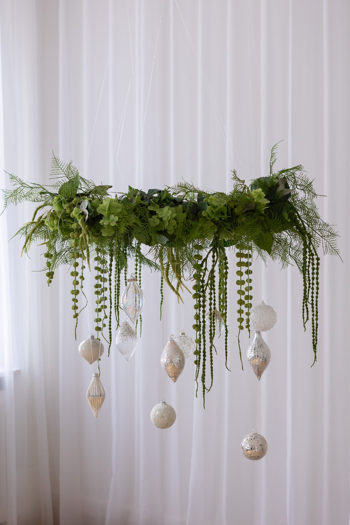 Stor krans af grønne kunstige blomster med hængende julekugler i hvide og sølv nuancer