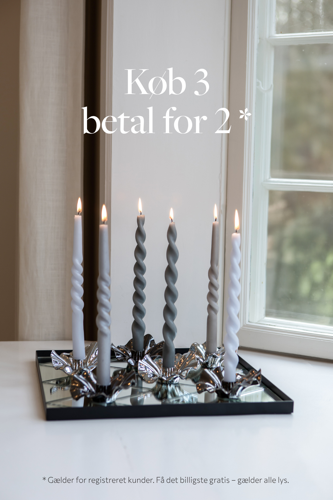 Fem snoede lys i farverne hvid og grå placeret i sølvfarvede lysestager på et spejlende fad. Lysene er tændt og står foran et vindue. Øverst på billedet står teksten 'Køb 3 betal for 2*'. Nederst på billedet står teksten 'Gælder for registreret kunder. Få det billigste gratis – gælder alle lys.