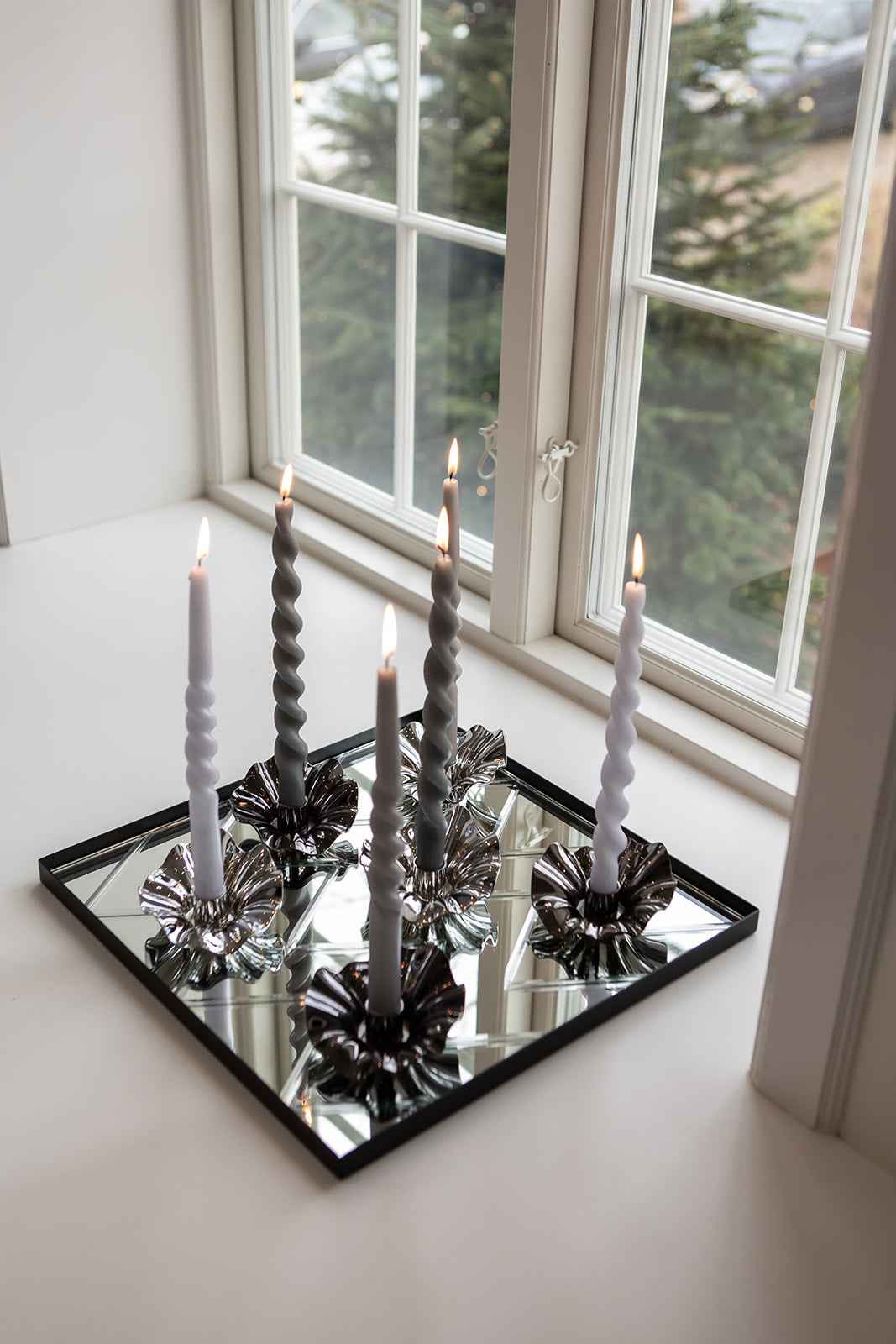 Fem snoede lys i grå og hvide nuancer placeret i sølvfarvede blomsterformede lysestager, stående på en kvadratisk spejlbakke foran et vindue.