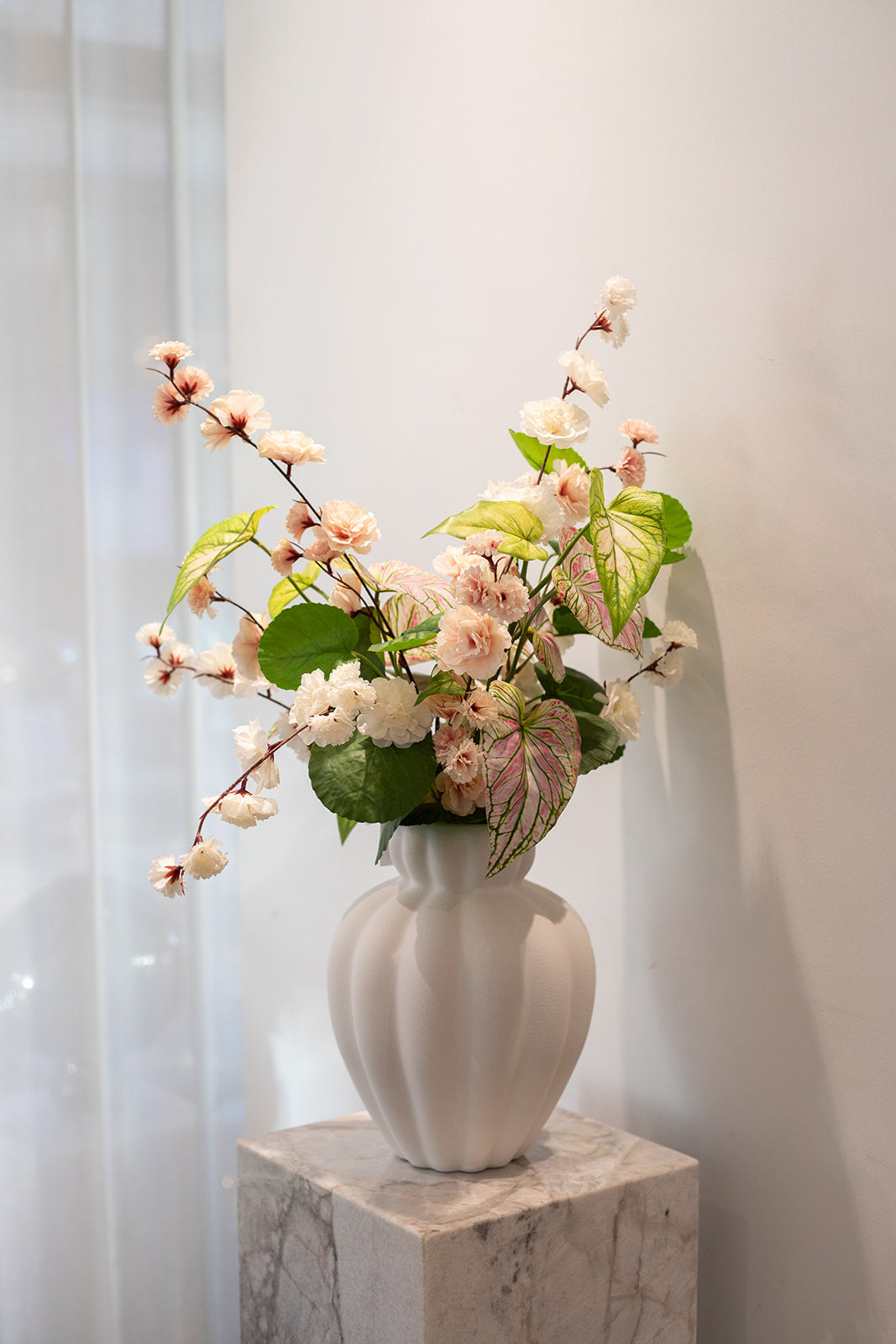 Kunstig buket i sarte rosa, hvide og grønne nuancer. Placeret i en hvid keramik vase, der står på en marmor pedestal. Baggrunden er hvid.