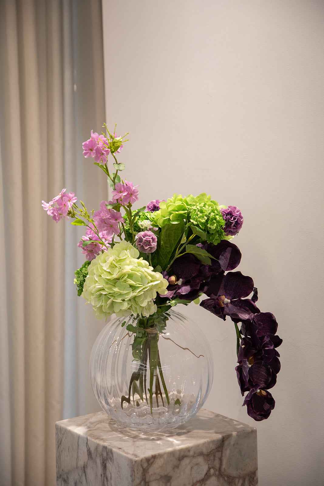 Buket af kunstige blomster i lilla og grønne nuancer. Placeret i en gennemsigtig glasvase, der står oven på en marmor pedestal.