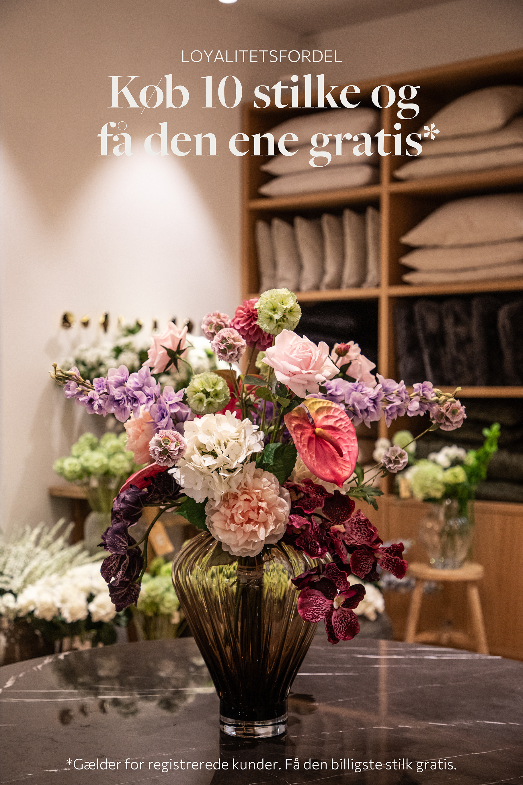 Stor farverig buket af kunstige blomster, placeret i en stor glasvase, der står på et marmor bord.