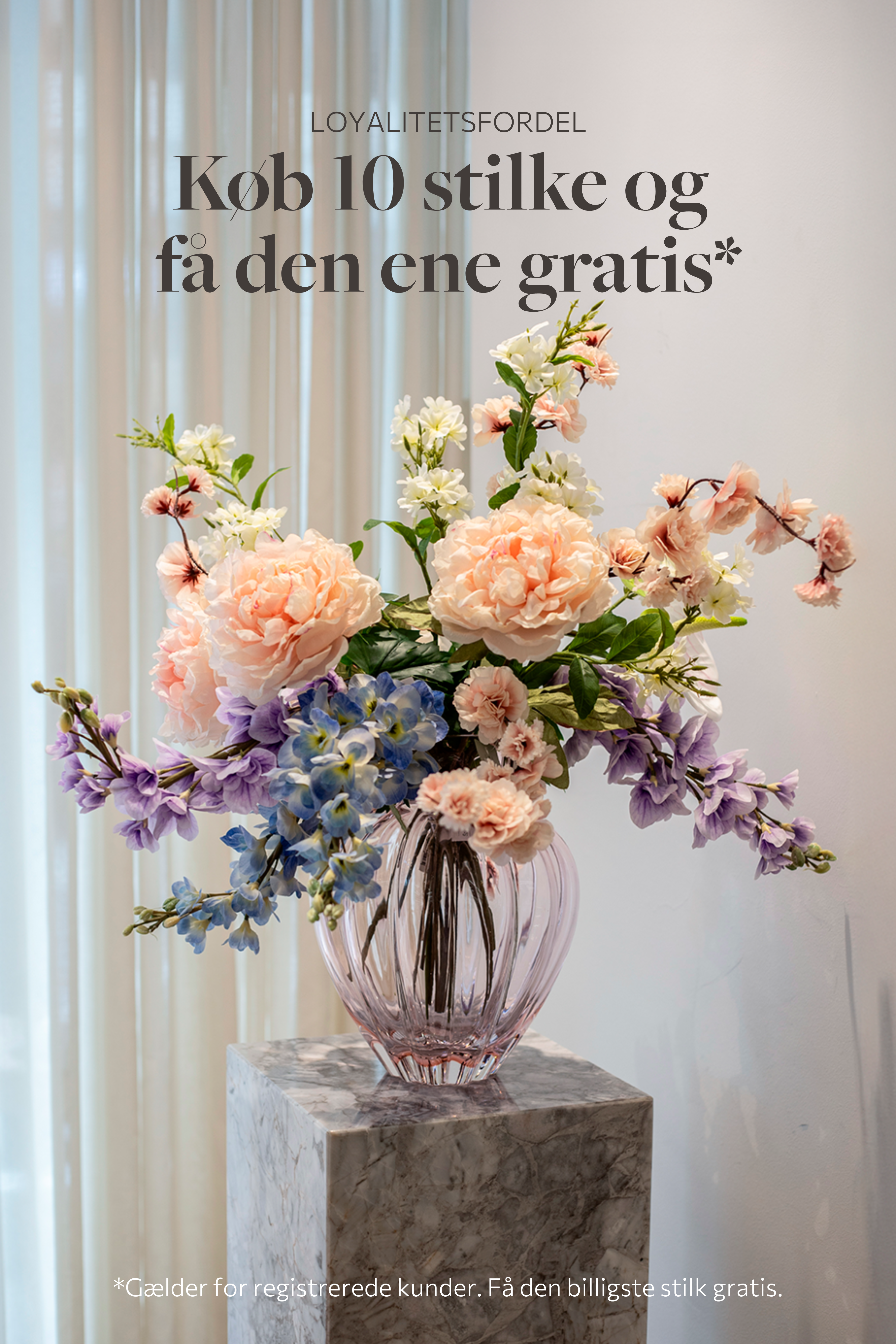 Stor farverig buket i rose, lilla, blå og hvide nuancer. Placeret i en lyserød glasvase, der står på en marmor pedestal.