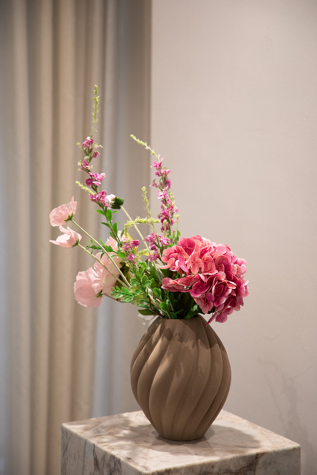 Buket af kunstige blomster i lyserøde og grønne nuancer. Placeret i en brun keramikvase, der står på en marmor pedestal.