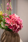 Nærbillede af en lyserød hortensia, der er en del af buket af kunstige blomster i lyserøde og grønne nuancer. Placeret i en brun keramikvase.