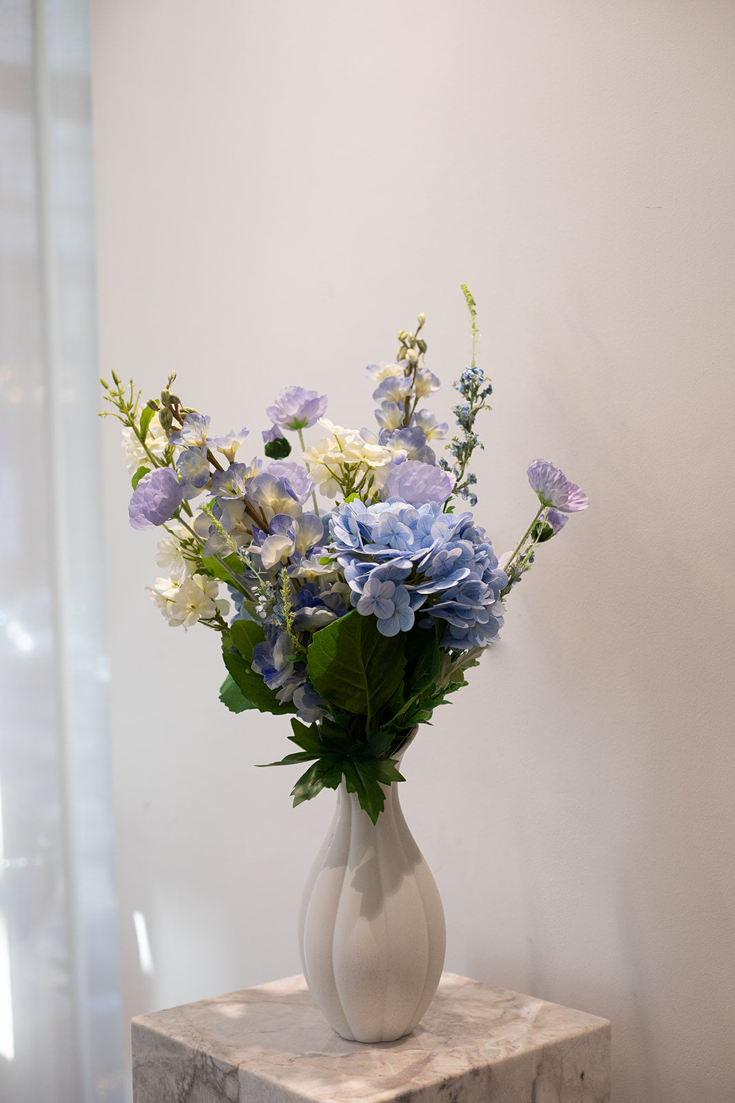 Kunstig buket i blå, lilla, hvide og grønne nuancer, placeret i en hvid keramik vase, der står på en marmor pedestal.