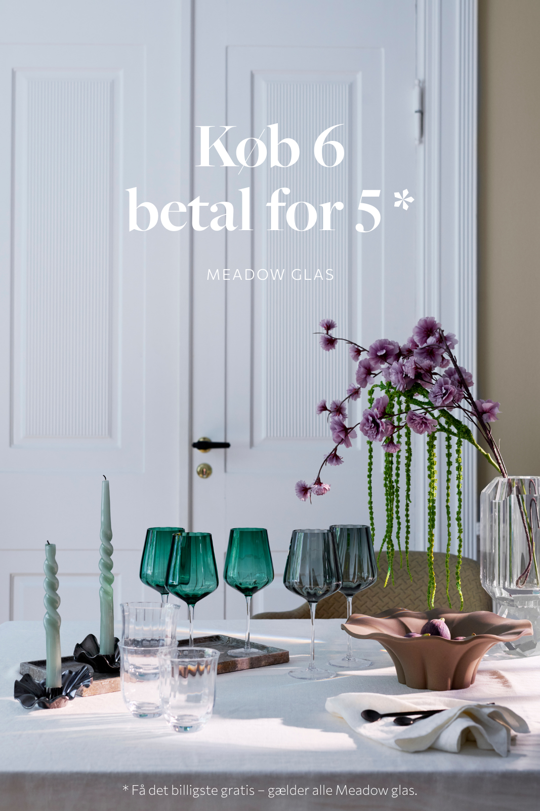 Kampagnebillede med teksten "Køb 6 betal for 5 – Meadow Glas". På bordet står fire Meadow vinglas, to i grøn glasfarve og to i røggrå. Glassene er placeret foran en hvid dobbeltdør. På bordet ses også snoede grønne lys i sorte stager, to klare vandglas, en brun skål med dekorative elementer samt en stor glasvase med lilla blomster og grønne hængende grene. Nederst i billedet står der: "Få det billigste gratis – gælder alle Meadow glas".