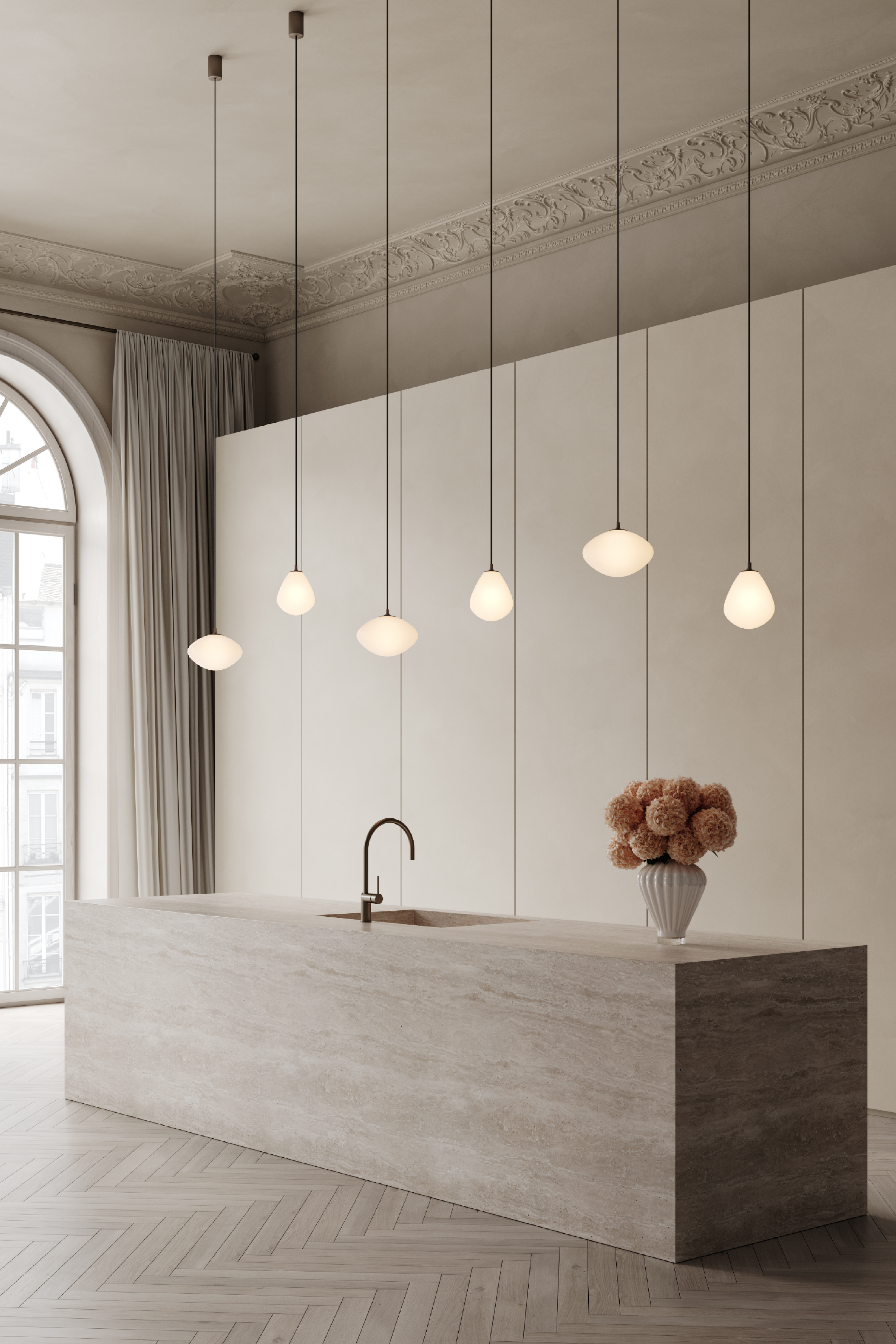 Et moderne køkken med en stor køkkenø i lys natursten og indbygget vask med buet armatur i messing. Over øen hænger seks slanke pendellamper med hvide glasskærme i forskellige organiske former. Til højre på bordpladen står en hvid riflet vase med tørrede blomster. Baggrunden består af høje, glatte vægpaneler, og til venstre ses et stort buet vindue med lyse gardiner. Gulvet er lagt i sildebensmønster.