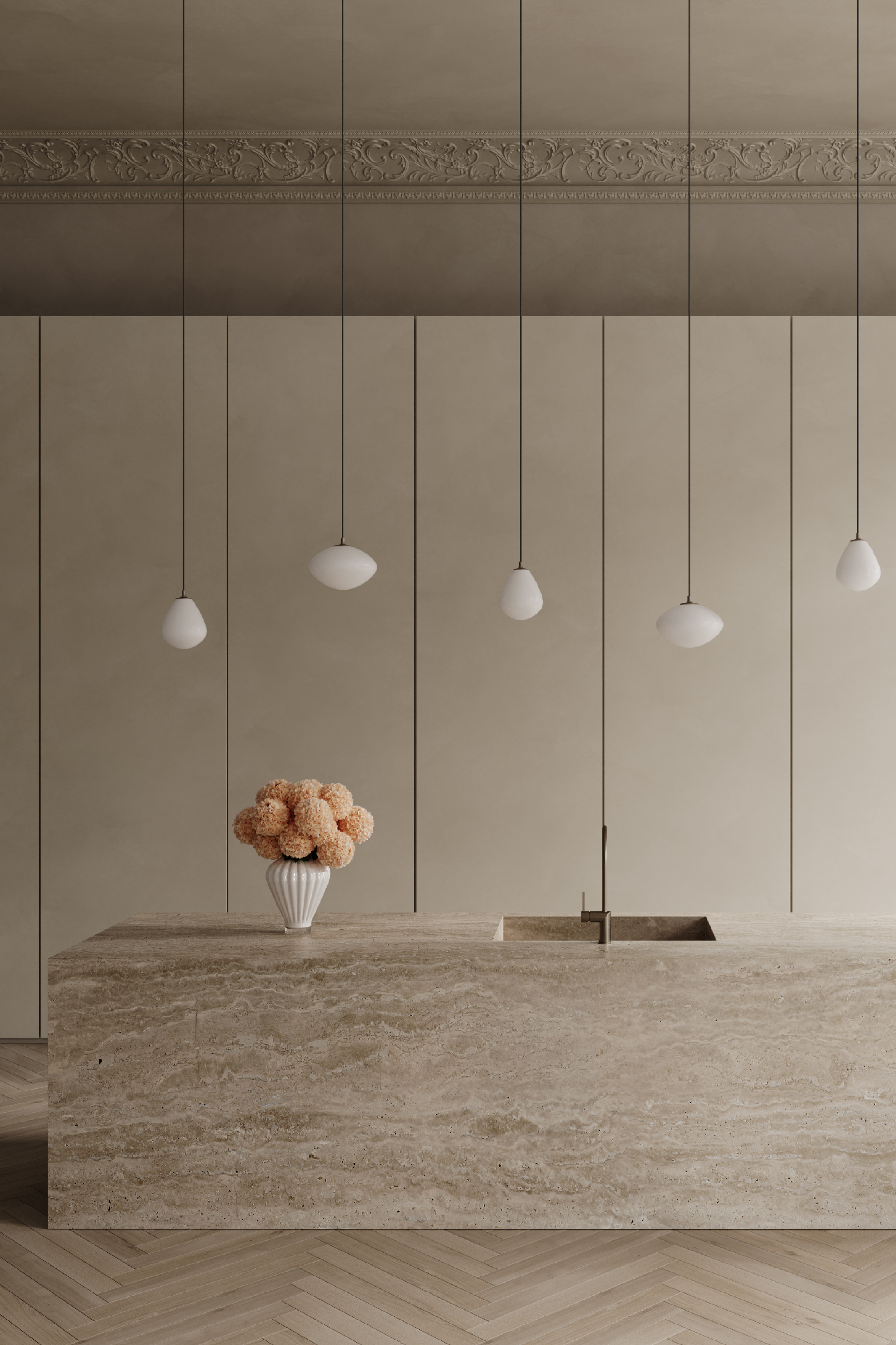 Et moderne køkken med en stor køkkenø i natursten med indbygget vask og slankt armatur i messing. Over øen hænger fem pendellamper med hvide, dråbeformede og ovale glasskærme. På bordpladen står en hvid riflet vase med en buket lyserøde blomster. Baggrunden består af høje vægpaneler i beige nuancer, og gulvet er lagt i sildebensmønster.