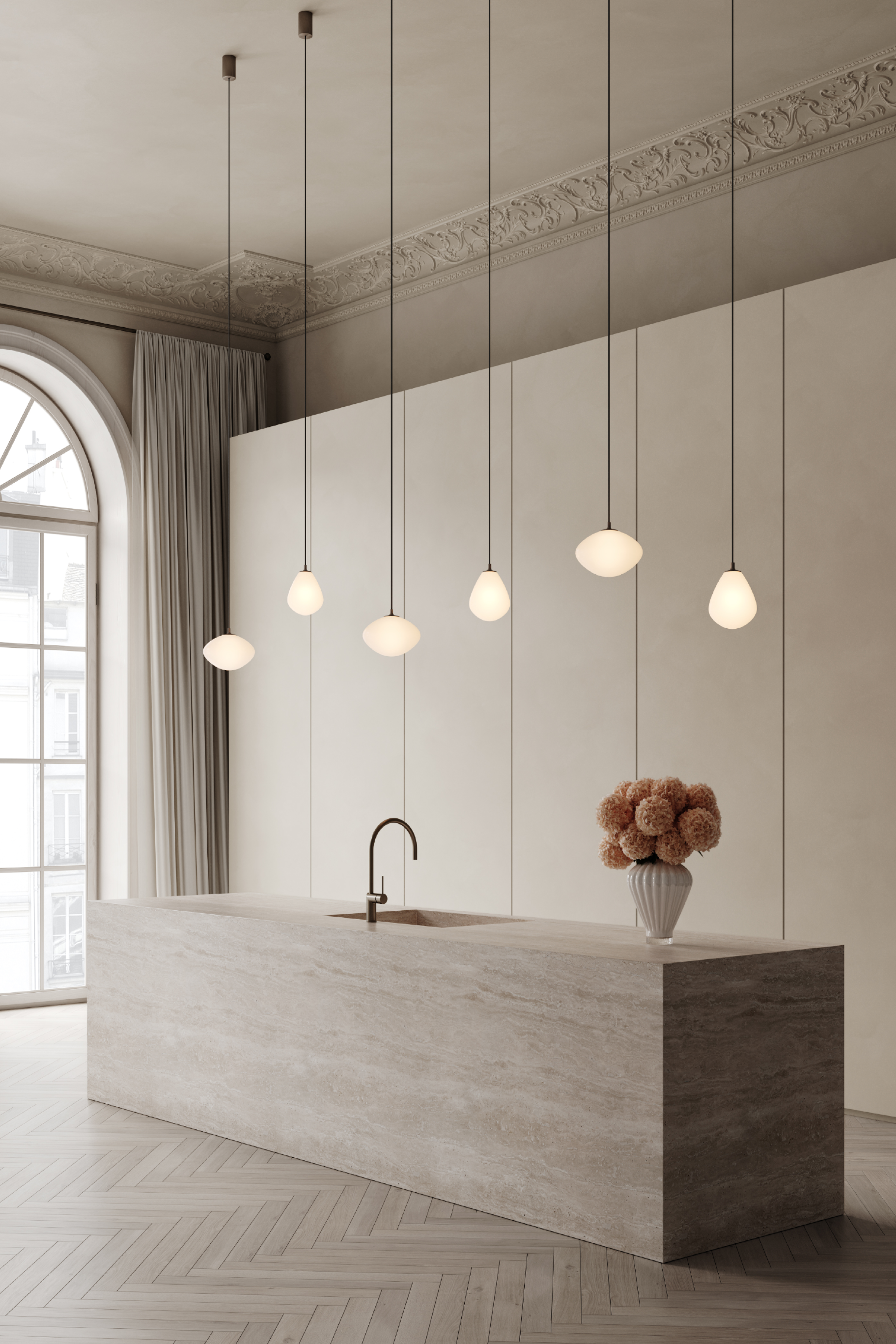 Et moderne køkken med en stor køkkenø i lys natursten og indbygget vask med buet armatur i messing. Over øen hænger seks slanke pendellamper med hvide glasskærme i forskellige organiske former. Til højre på bordpladen står en hvid riflet vase med tørrede blomster. Baggrunden består af høje, glatte vægpaneler, og til venstre ses et stort buet vindue med lyse gardiner. Gulvet er lagt i sildebensmønster.