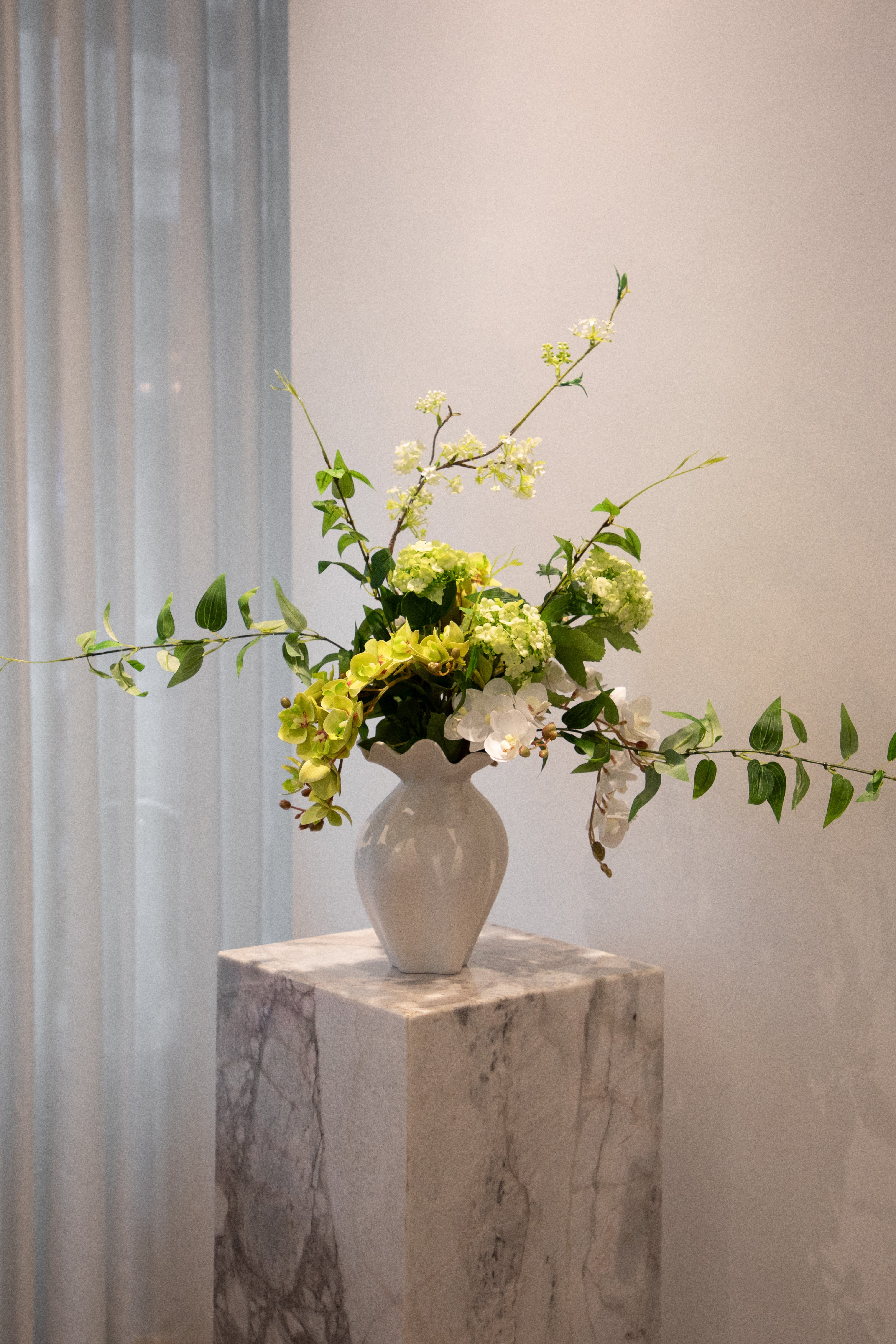 Kunstig buket i grønne og hvide nuancer, placeret i en hvid keramik vase, der står på en marmor pedestal.