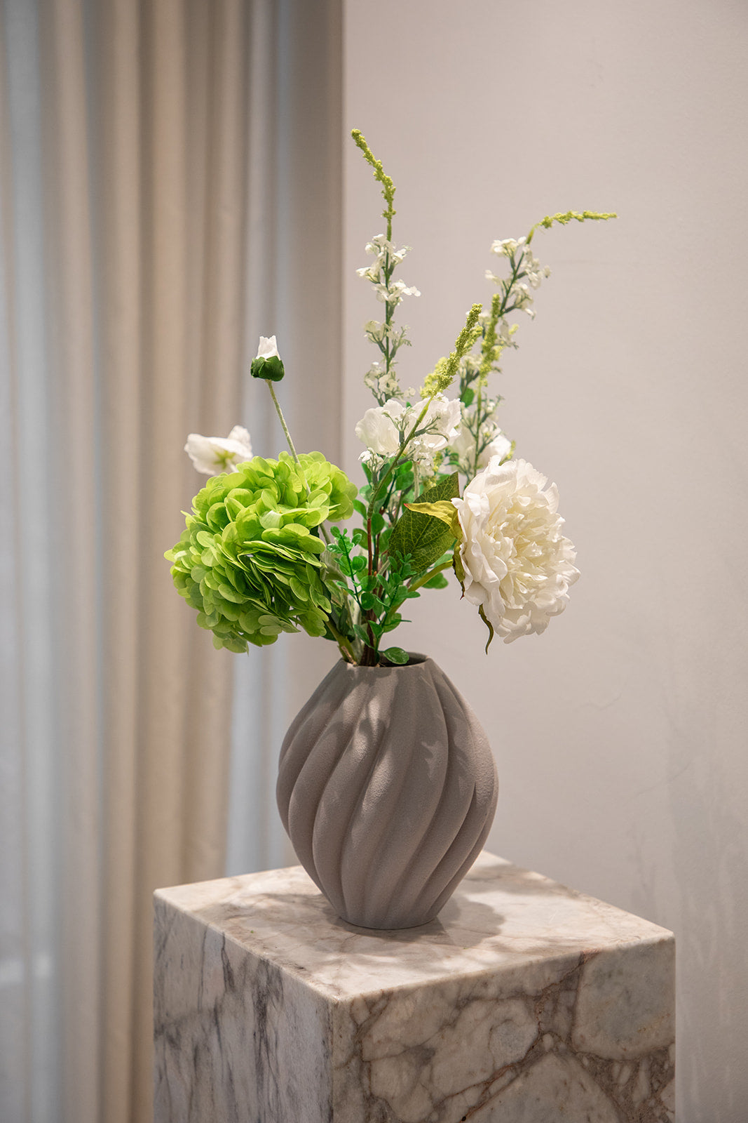 Buket af kunstige blomster i hvide og grønne nuancer. Placeret i en grå keramikvase, der står på en marmor pedestal.