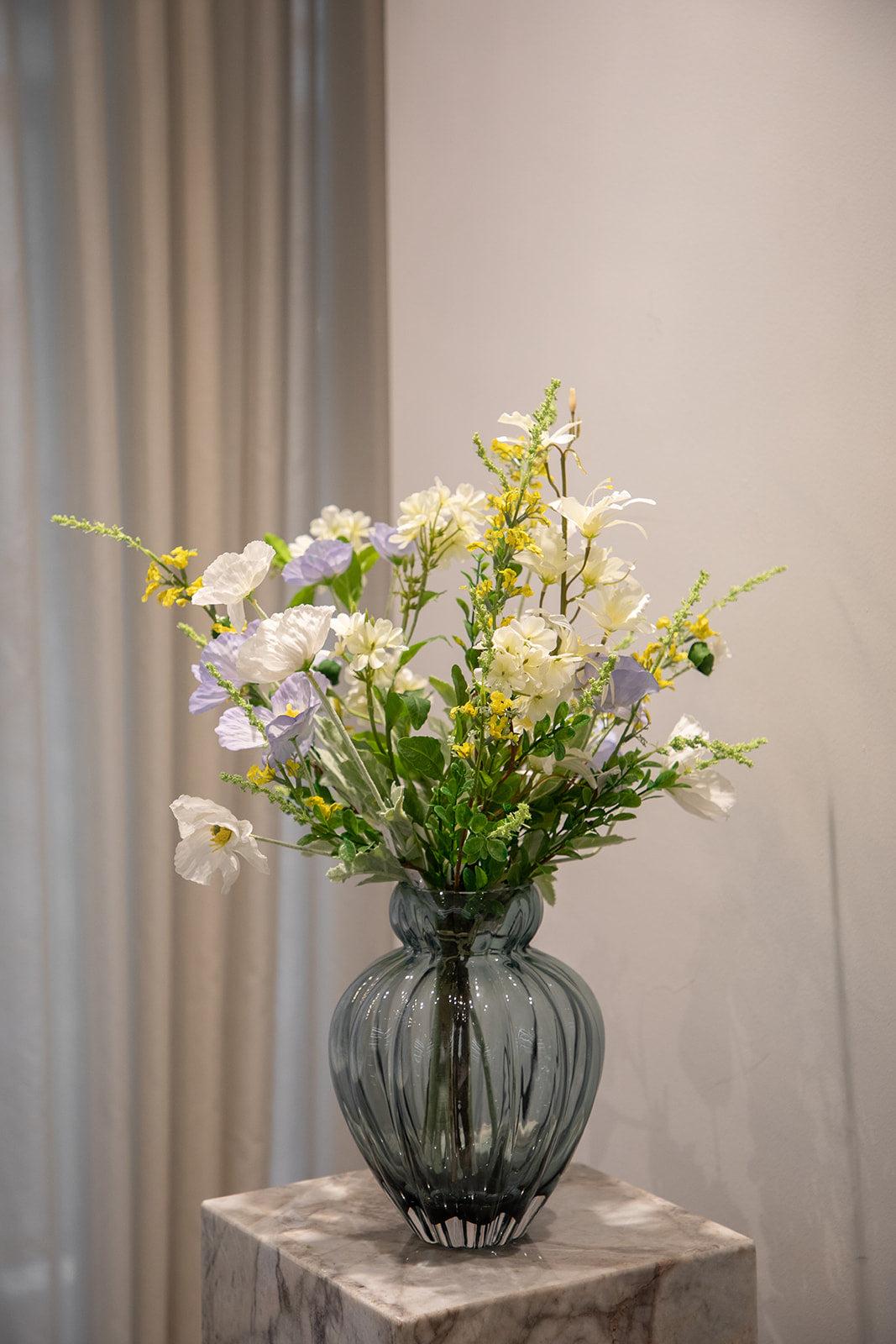 Buket af kunstige blomster i gule, hvide, lilla og grønne nuancer. Placeret i en grålig glasvase, der står på en marmor pedestal.