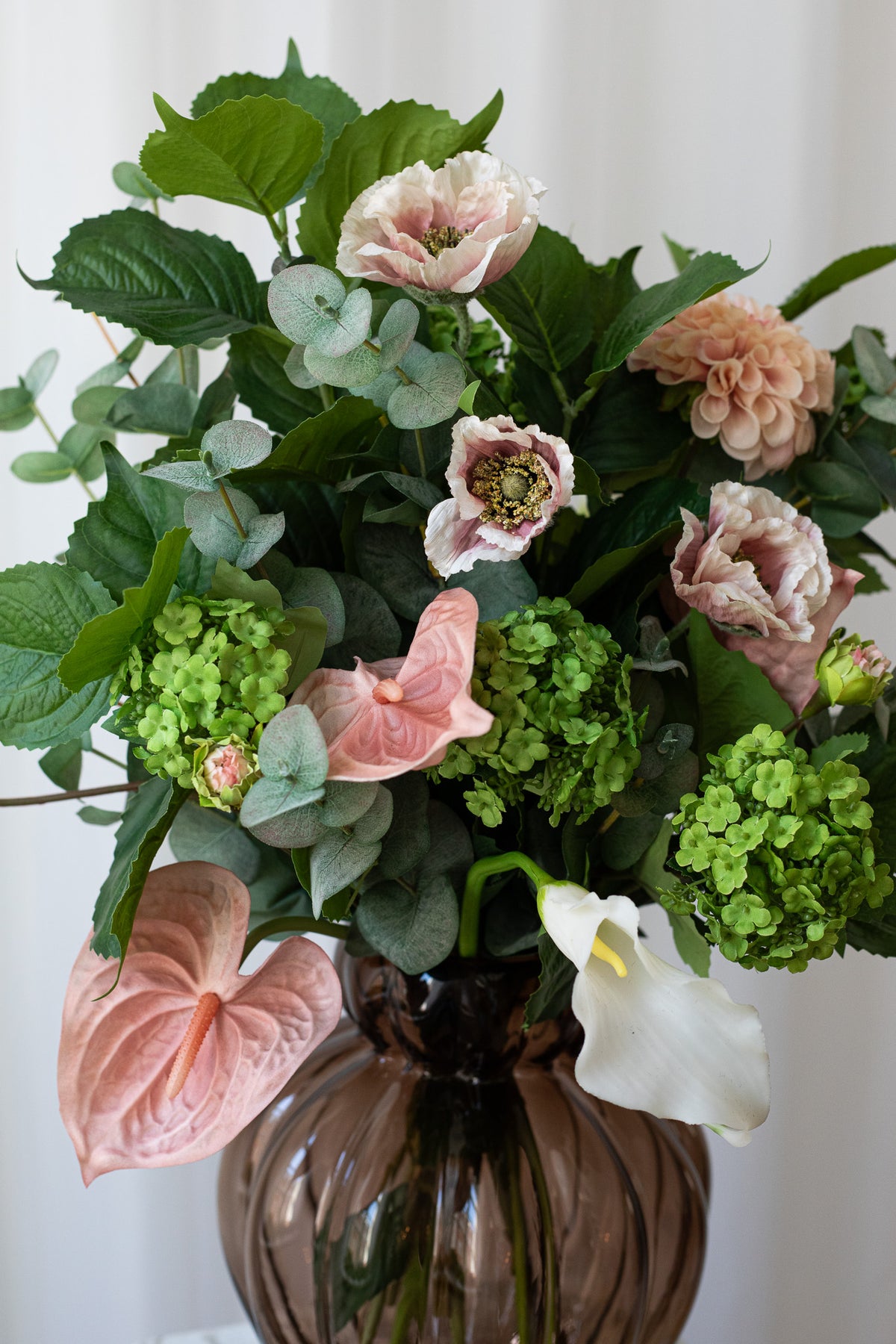 En kunstig buket med valmuer, flamingoblomst, calla liljer og eukalyptusgrene. Buketten har smukke grønne og lyserøde farver.