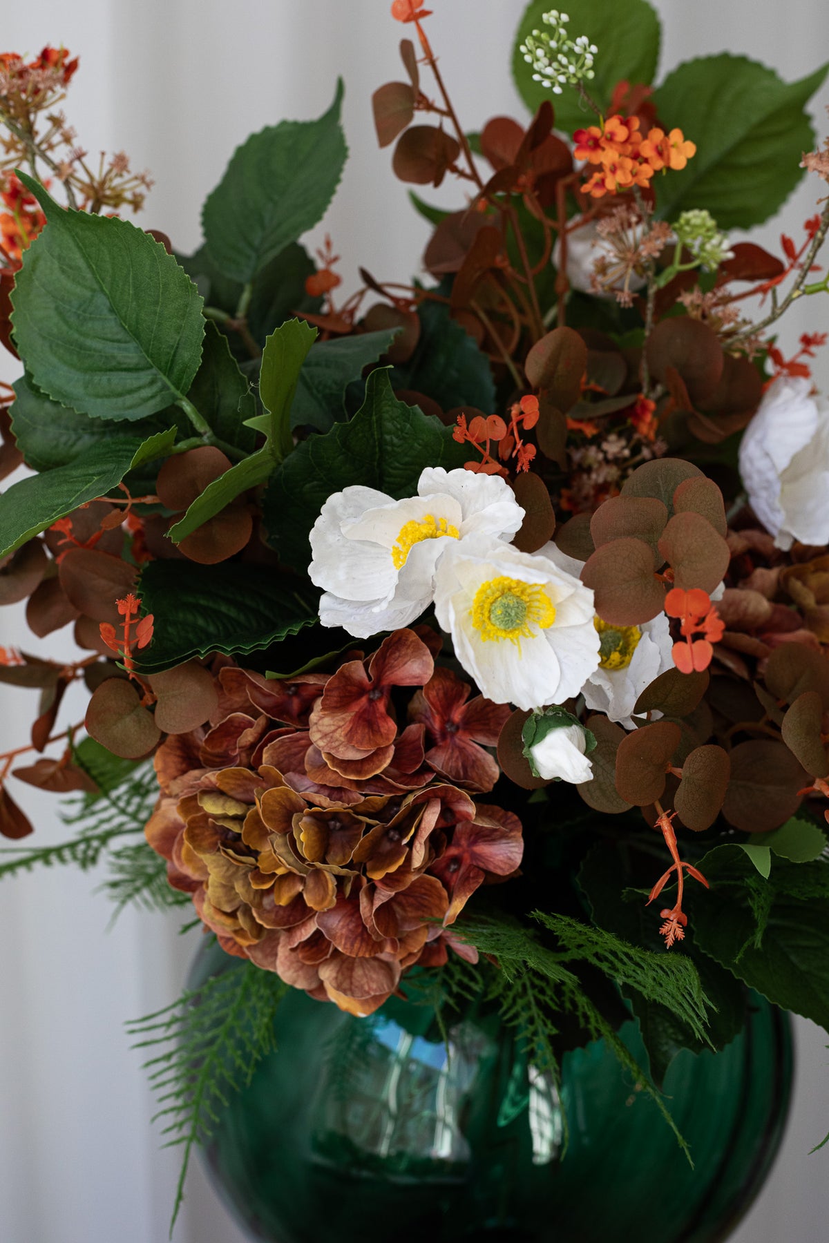En gylden buket i efterårets farver, der er sammensat af hvide valmuer, en orange hortensia, grønne og orange bladgrene.