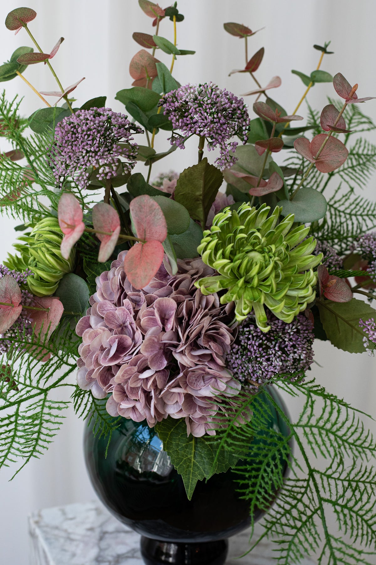 Nærbillede af en grøn og lyselilla buket skabt af hortensia stilke, bærgrene, eukalyptusgrene og andet grønt. Den naturtro, kunstige buket er fra Specktrum.