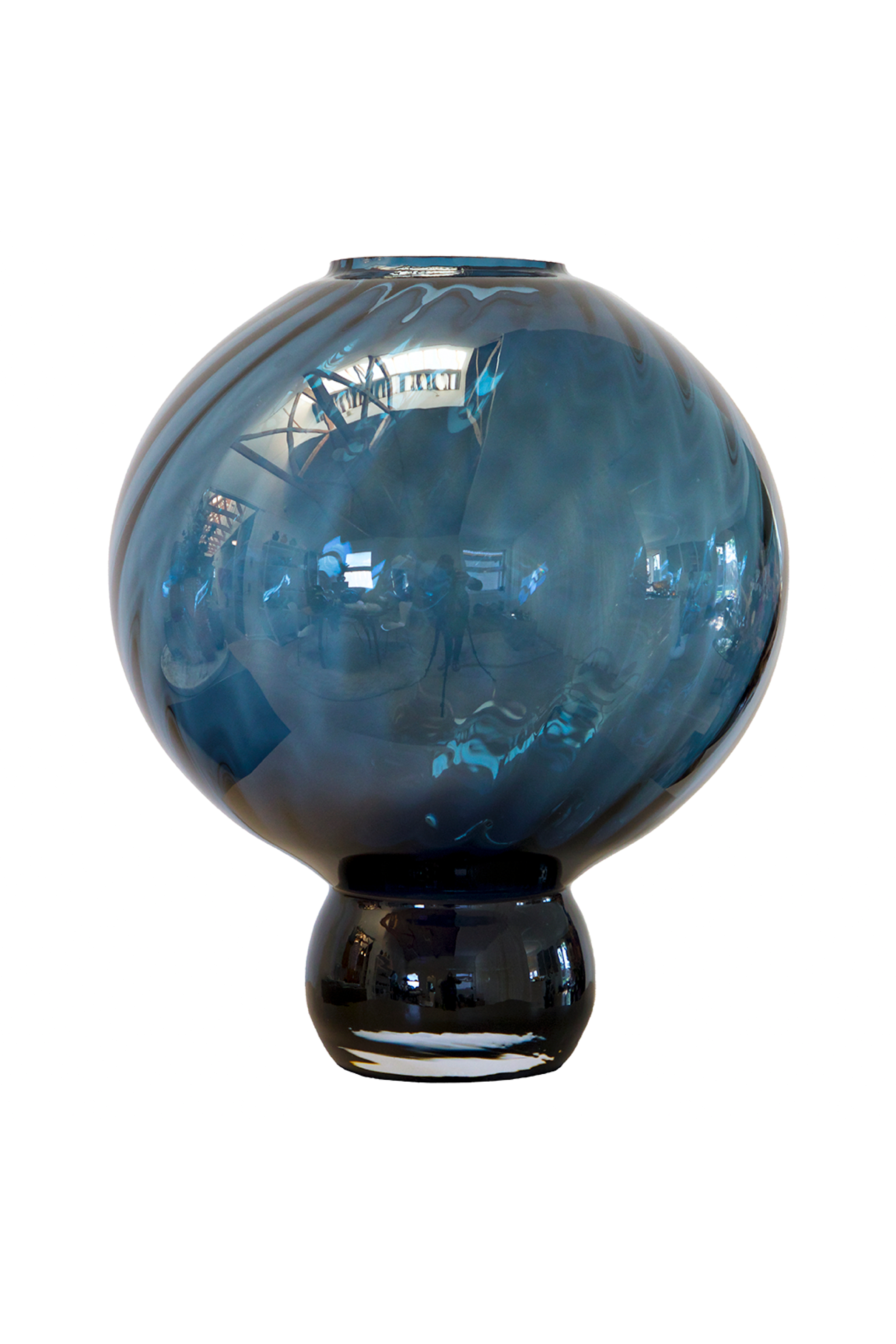 En rund blå glasvase med mørk fod og swirl-mønster i glasset. Vasens overflade er blank og reflekterer omgivelserne.