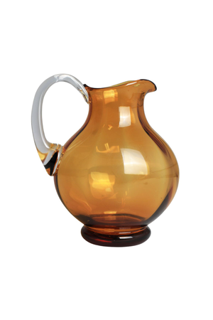 Ravfarvet glaskande med rund form og kort fod. Kanden har en klar glas-hank og en markeret hældetud. Produktet er fotograferet mod en hvid baggrund.