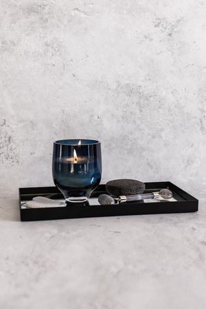 Et tændt duftlys i mørkeblåt glas står på et sort rektangulært fad med spejlbund. Ved siden af ligger en hvid gua sha-sten, en sort pumice-sten og en grå ansigtsrulle.