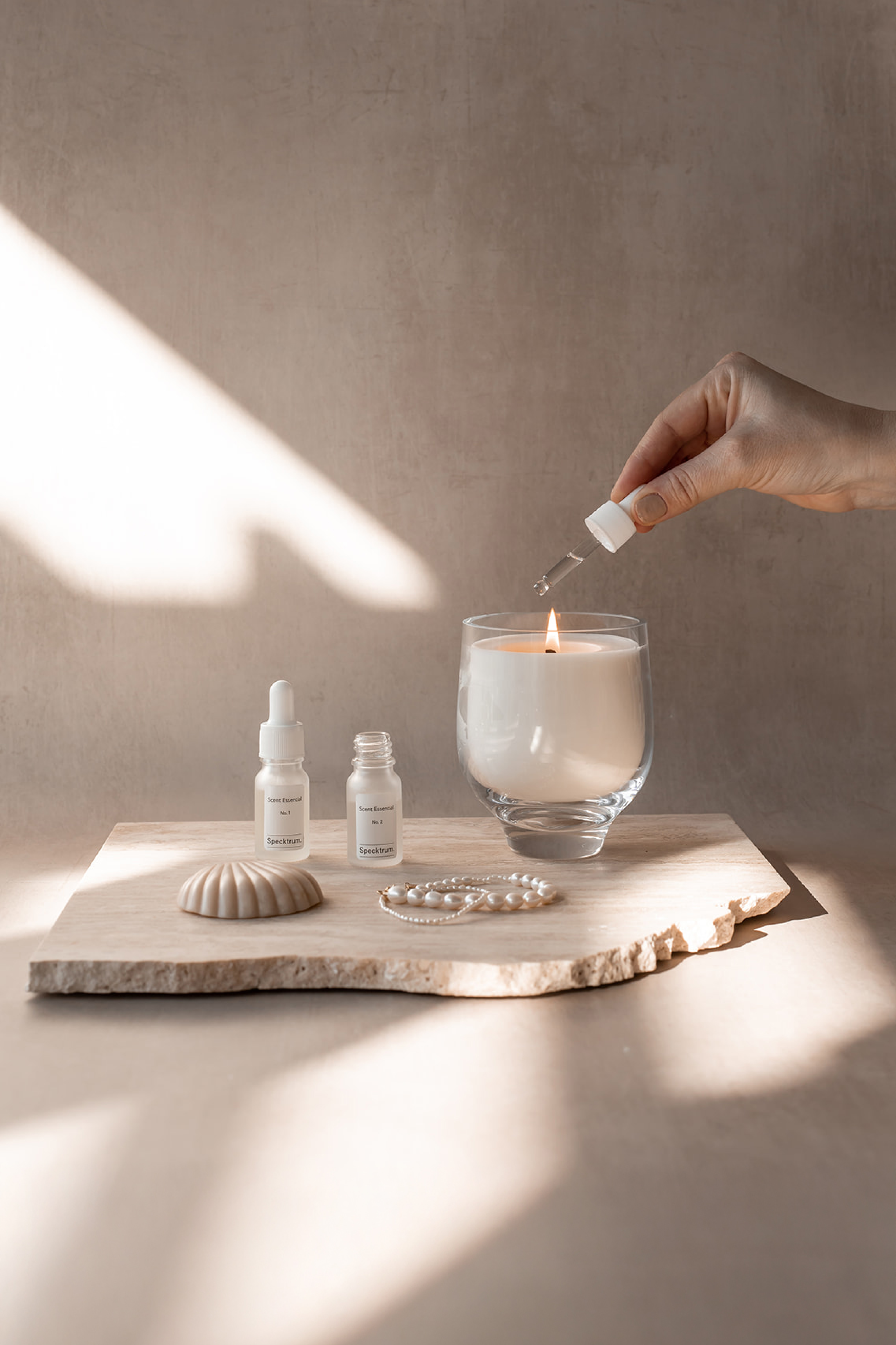 Et tændt hvidt duftlys i klar glasbeholder står på en lys stenplade sammen med to små pipetteflasker mærket 'Specktrum Scent Essentials', en dekorativ muslingeskal og en perlekæde. En hånd holder en pipette over lyset.