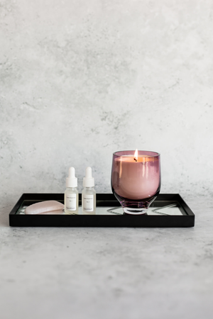 Et lilla glas med tændt lys placeret på en sort rektangulær bakke med spejlbund. På bakken står også to små flasker med pipette og hvide etiketter samt en lys sten.