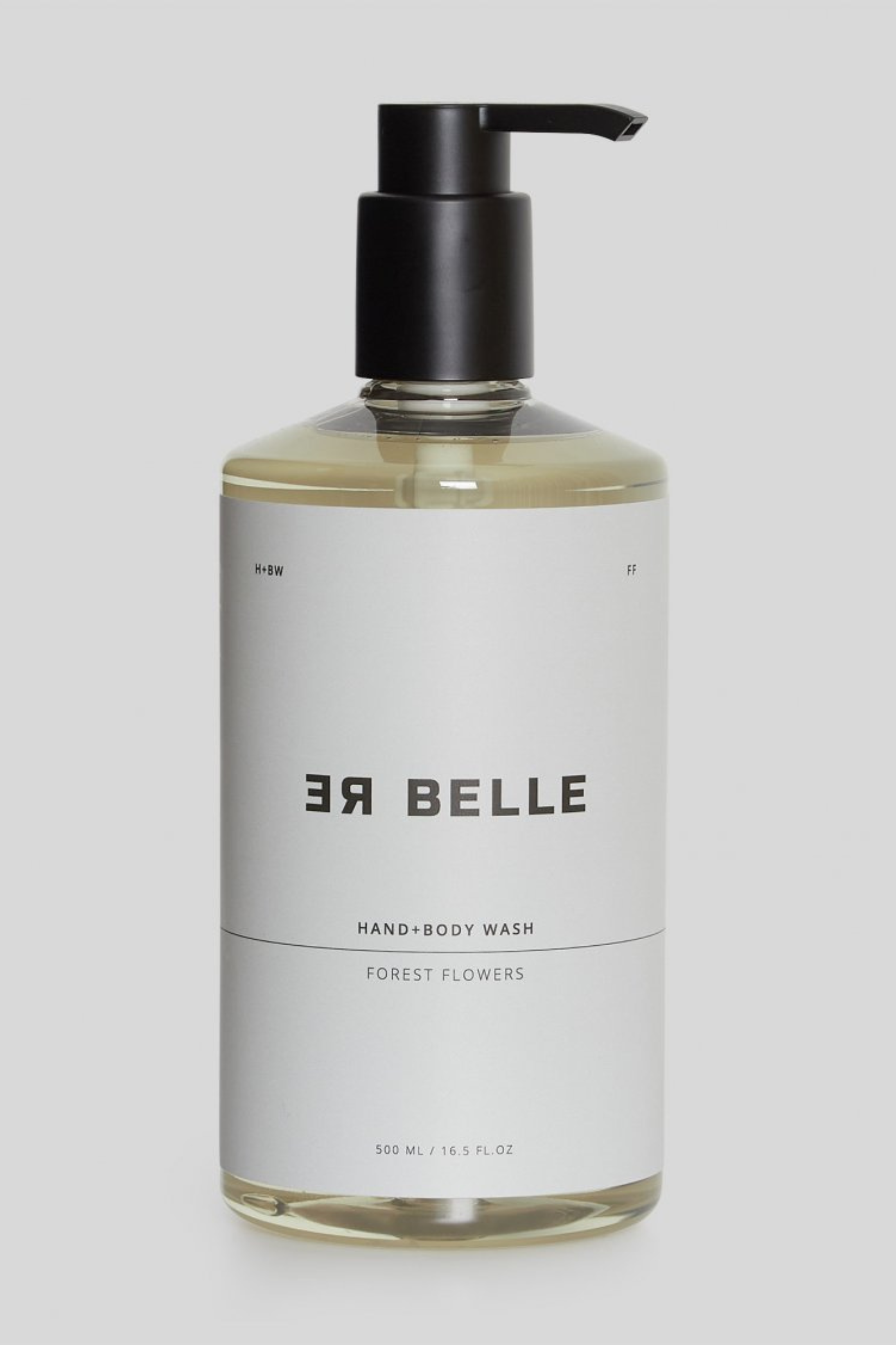 En gennemsigtig plastflaske med sort pumpe indeholdende en lys væske. Etiketten er hvid med sort tekst: “RE BELLE HAND+BODY WASH, FOREST FLOWERS, 500 ML