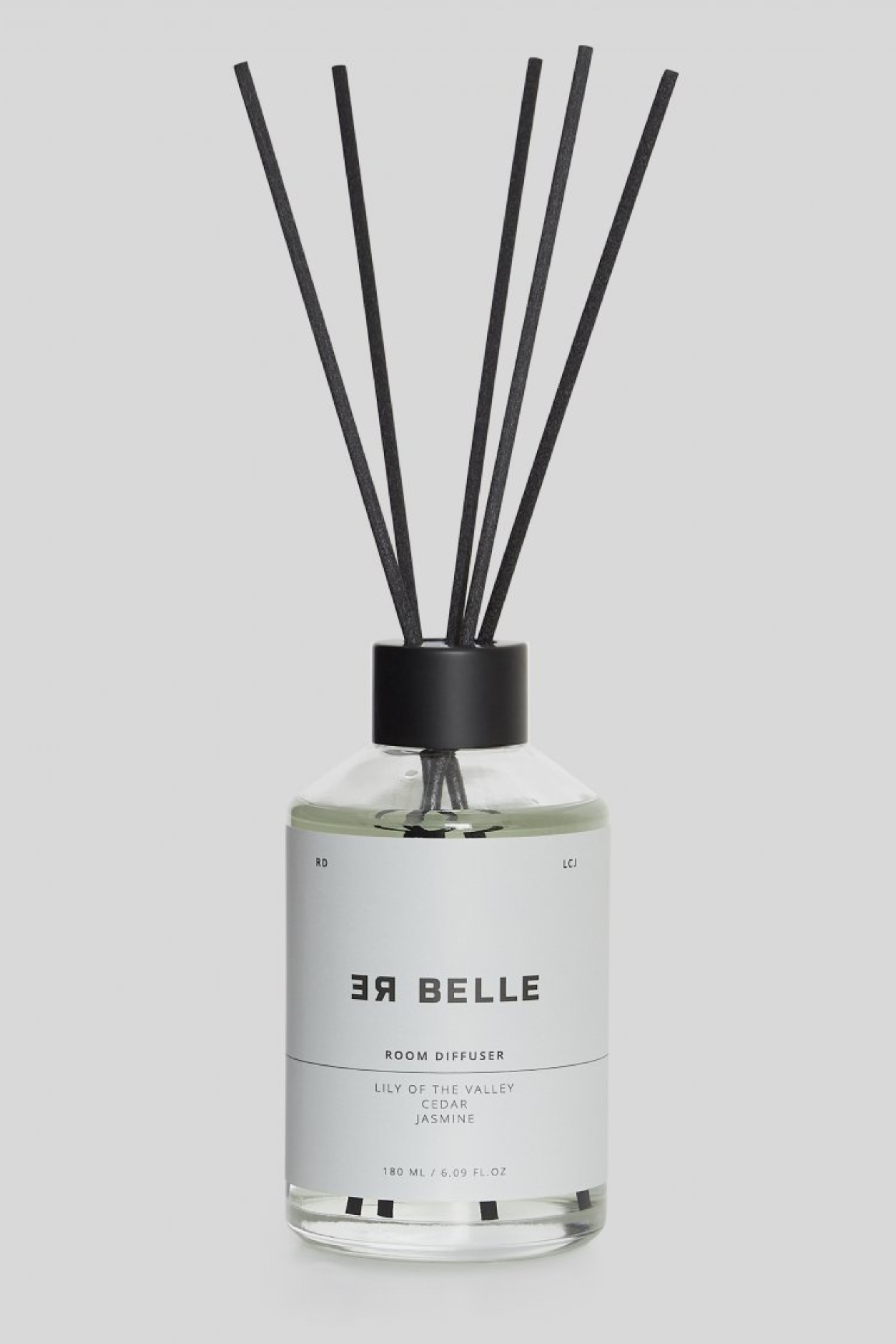En gennemsigtig glasflaske med sort låg og flere sorte duftpinde. Etiket i hvidt med sort tekst: “ER BELLE ROOM DIFFUSER, LILY OF THE VALLEY, CEDAR, JASMINE, 180 ML