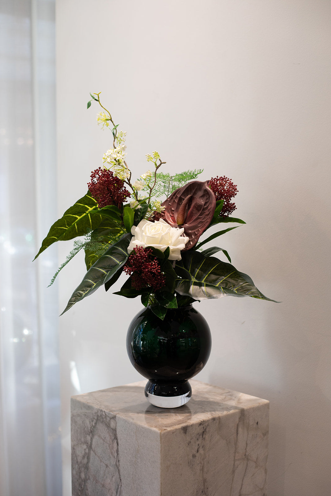 Kunstig buket i dybe røde, grønne og hvide nuancer. Placeret i en mørkegrøn glasvase, der står på en marmor pedestal.