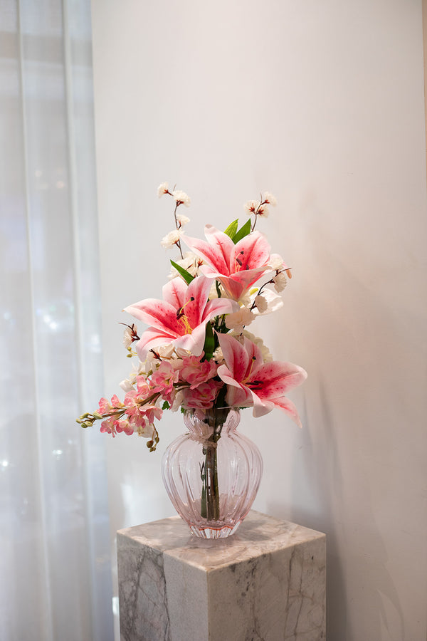 Kunstig buket blomster i lyserøde og creme nuancer. Placeret i en lyserød glas vase, der står på en marmor pedestal.