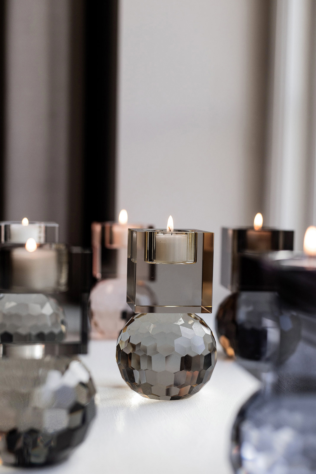 Flere tændte fyrfadslys i moderne lysestager af facetslebet krystalglas. Stagerne har kubeformet top og kugleformet base i forskellige farvetoner.