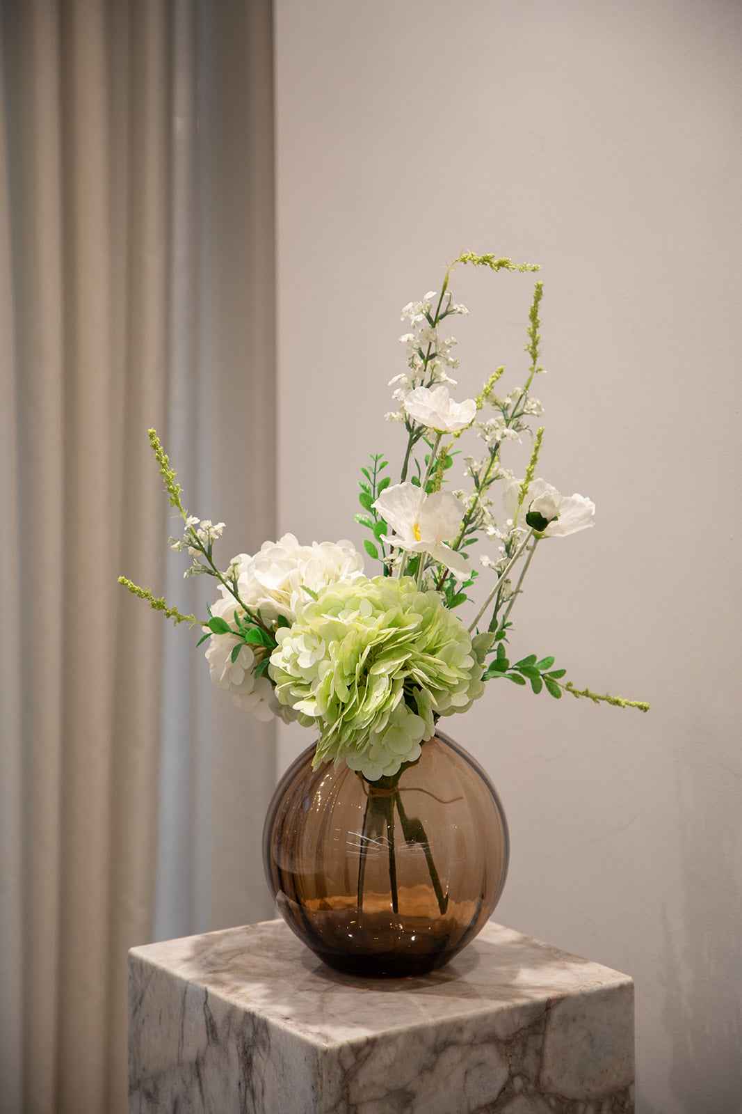 Stor buket af kunstige blomster i hvide og grønne nuancer. Placeret i en topaz farvede glasvase, der står oven på en marmor pedestal.