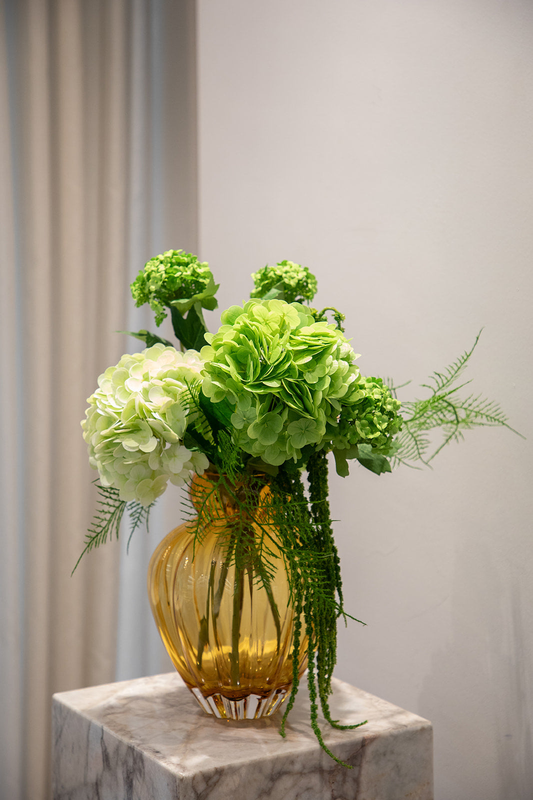 Buket af kunstige blomster i grønne nuancer. Placeret i en orange farvede glasvase, der står oven på en marmorpedestal.