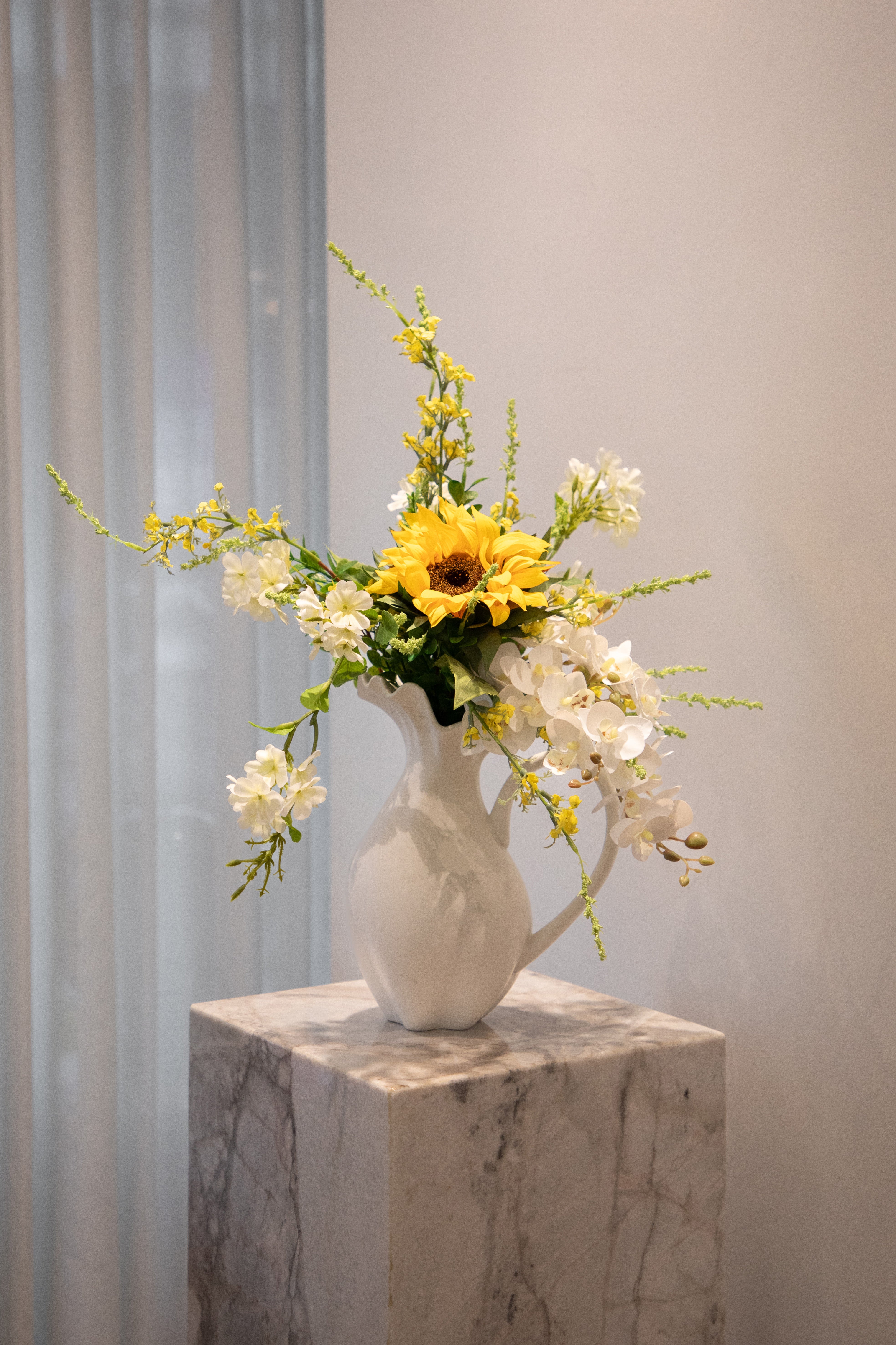 Kunstig buket i gule, hvide og grønne nuancer, placeret i en hvid keramik vase, der står på en marmor pedestal.