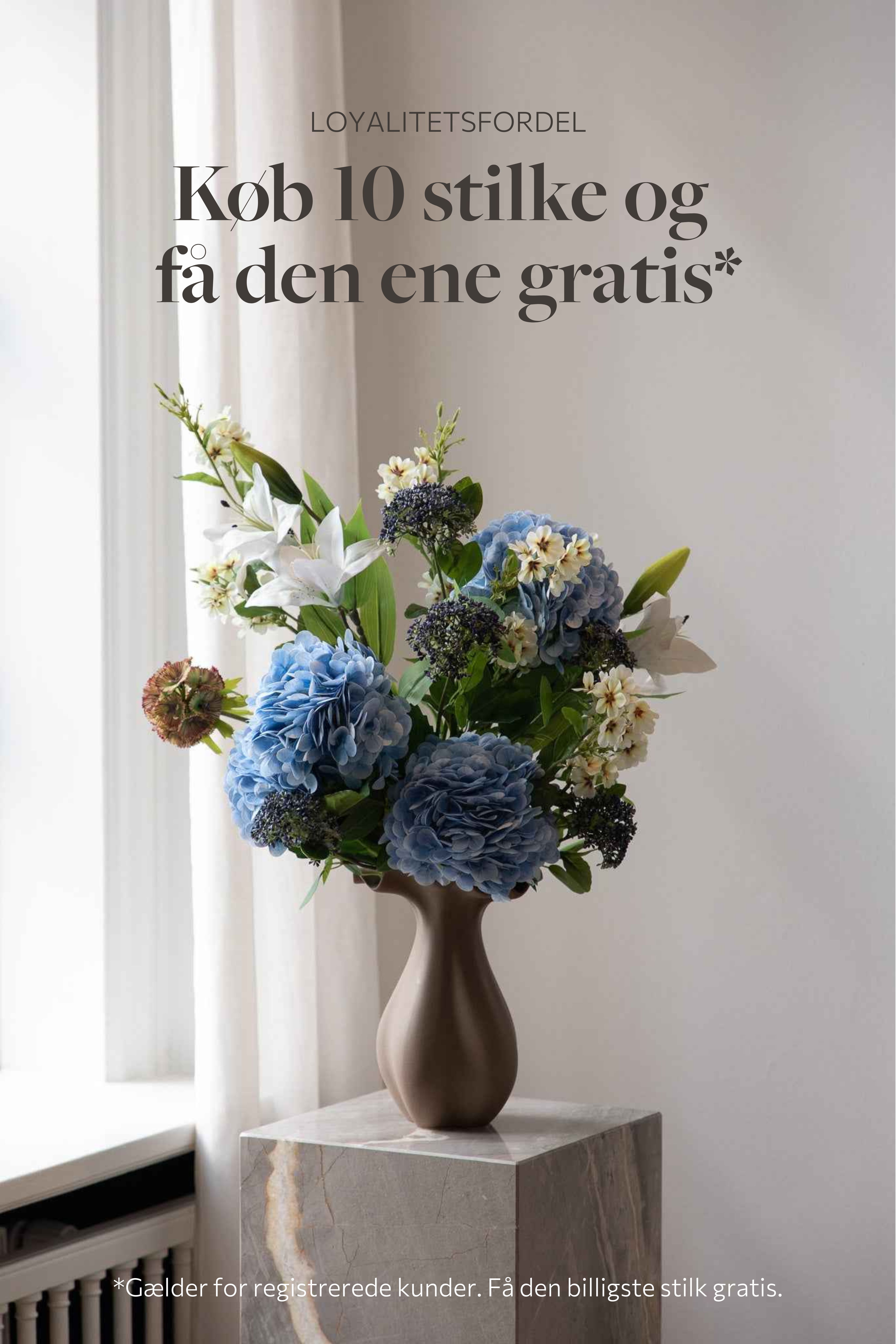 Farverig buket i blå, grønne og hvide nuancer af kunstige blomster. Placeret i en brun nellie keramikvase, står ovenpå en marmorpedestal.