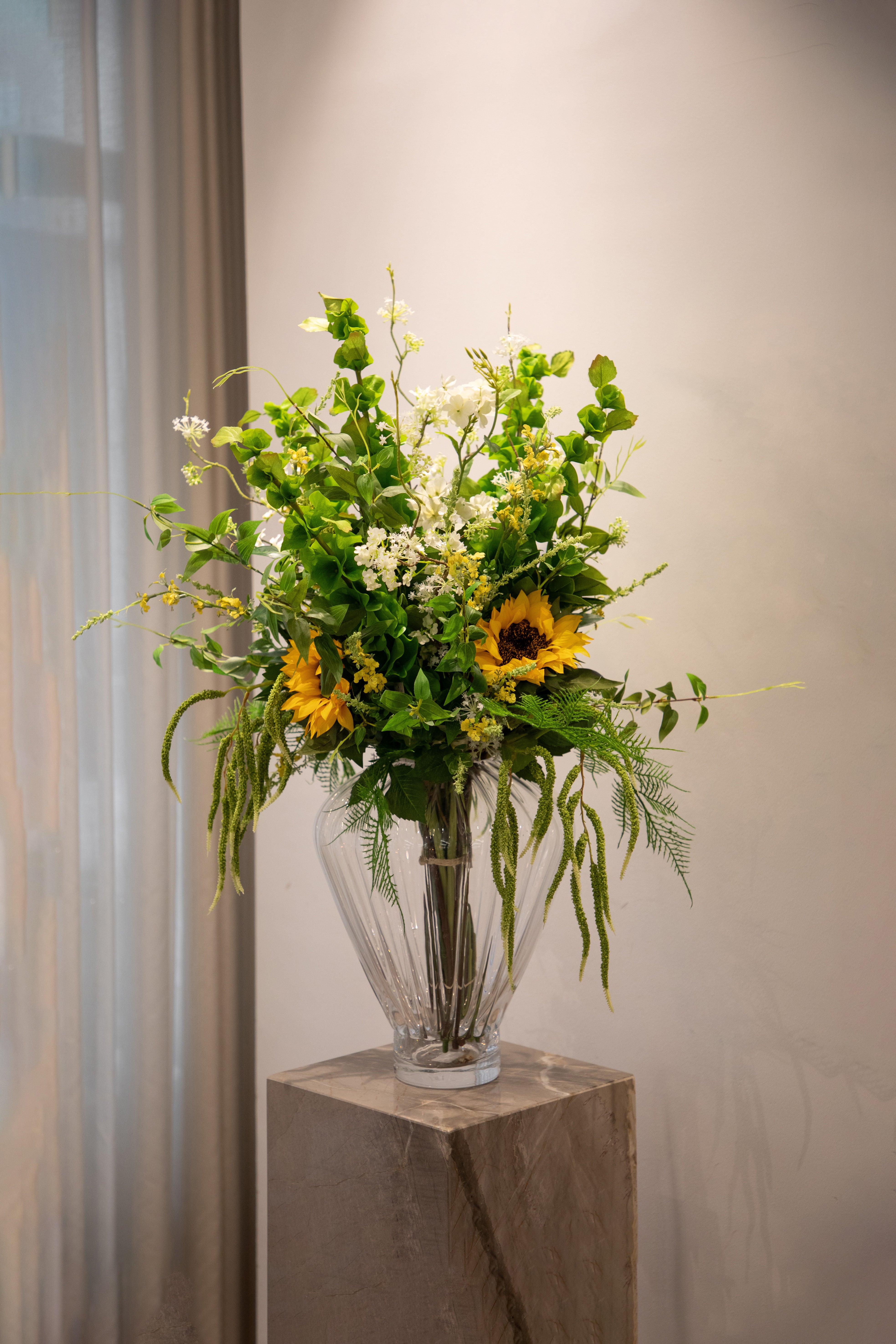 Stor buket af kunstige blomster i grønne, sarte hvide og gule nuancer. Buketten står i en Evelyn gennemsigtig glas vase fra Specktrum ovenpå en marmor pedestal.