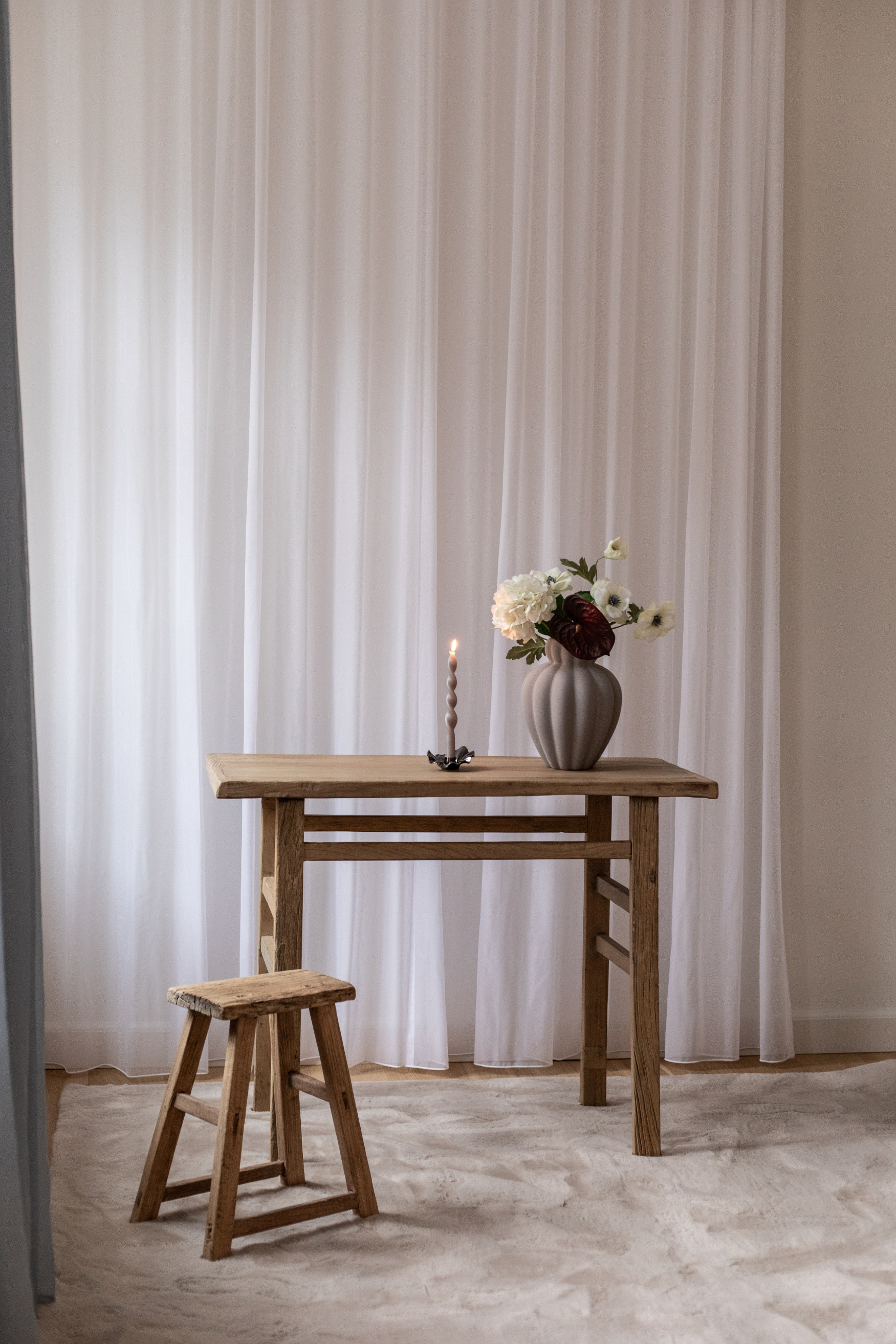 Et rustikt træbord står foran hvide gardiner med en lille træskammel ved siden af. På bordet står en grå vase med blomster og en tændt snoet lysestage.