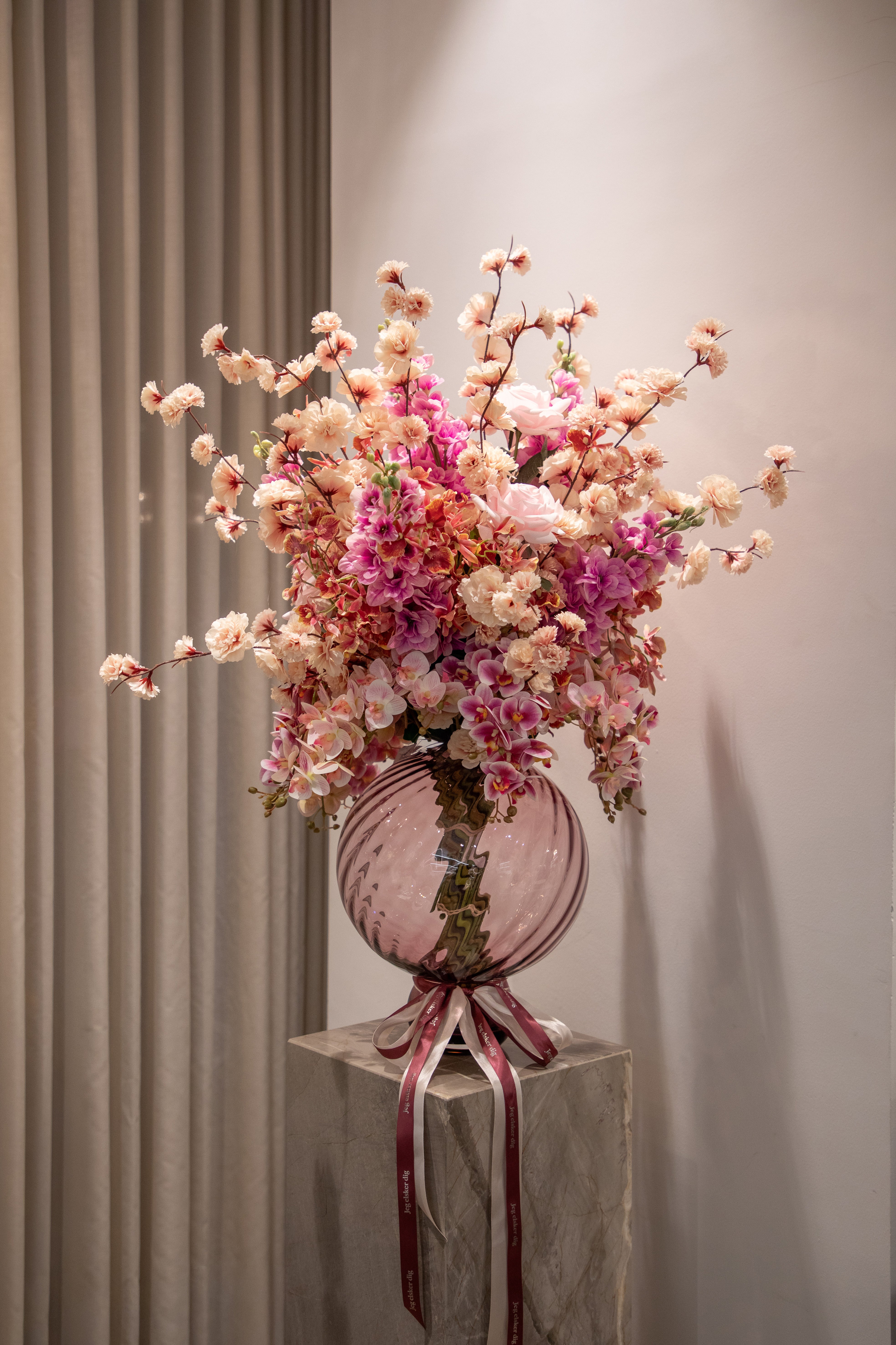 Valentines buket af kunstige blomster i rosa, lyserøde og creme farver. Buketten er placeret en i en rosa glas vase, der står på en marmor pedestal.