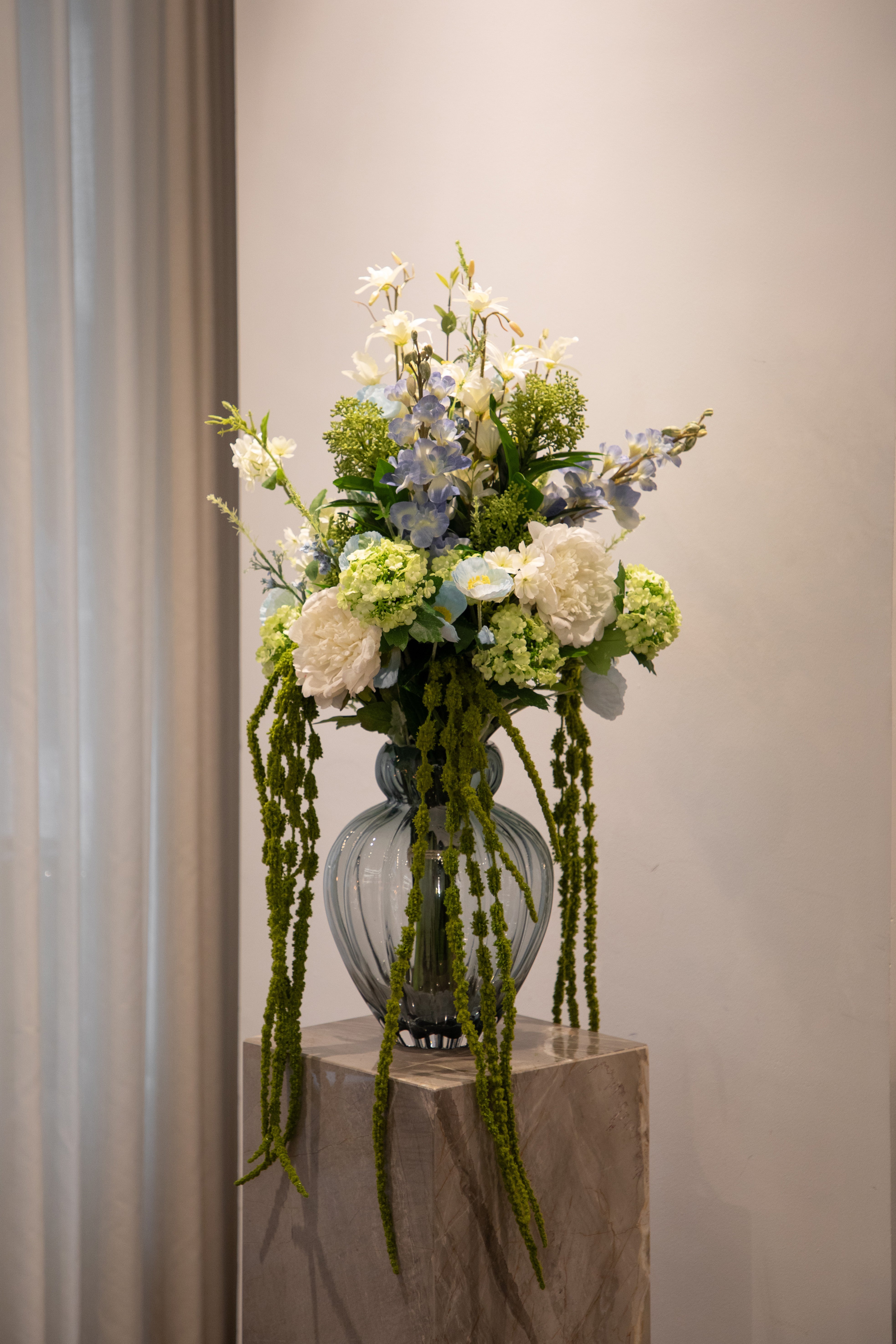 Buket af kunstige blomster i blå, hvide og grønne nuancer. Buketten står i en lyseblå/grå glasvase fra Specktrum ovenpå en marmor pedestal.