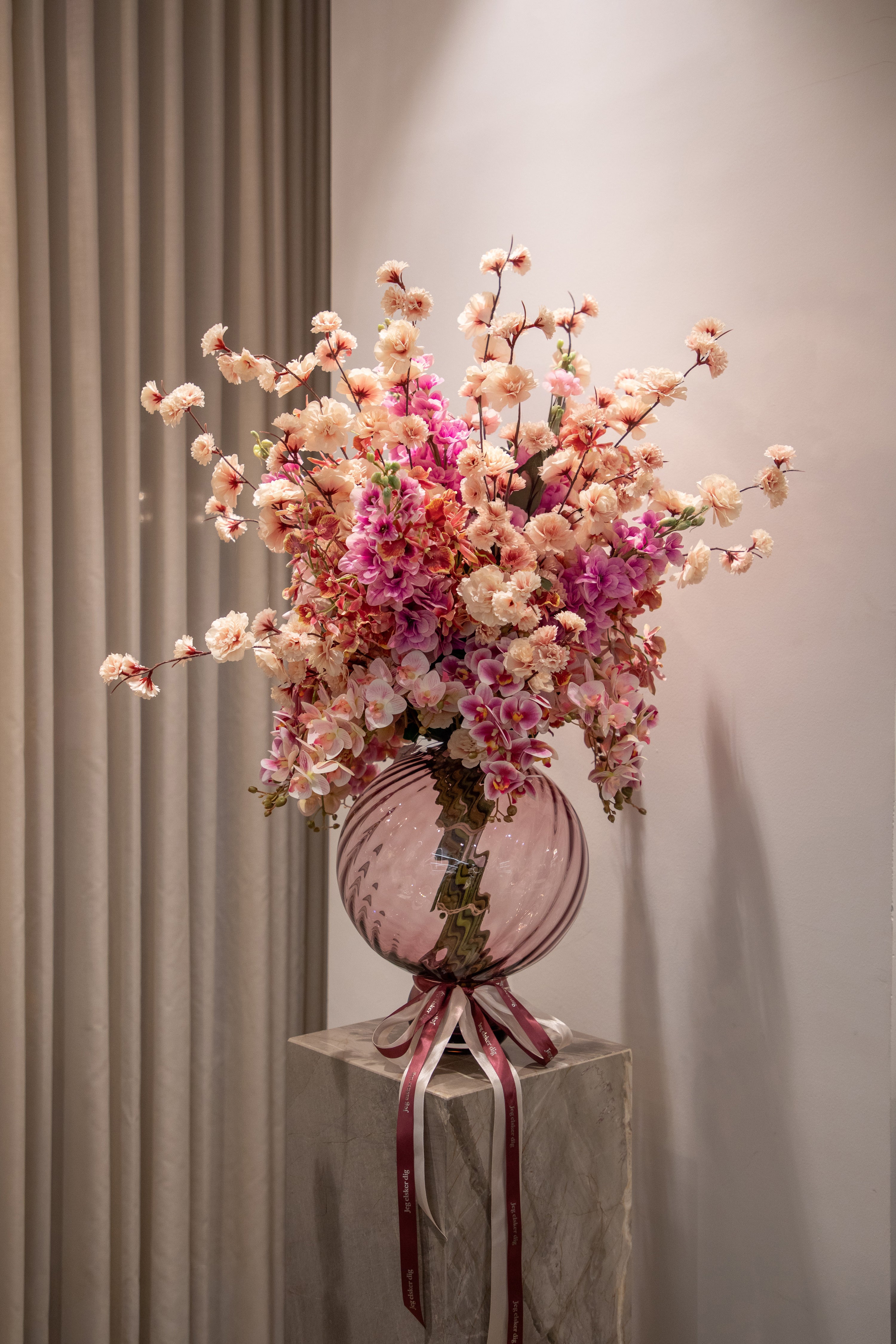 Valentines buket af kunstige blomster i rosa, lyserøde og creme farver. Buketten er placeret en i en rosa glas vase, der står på en marmor pedestal.