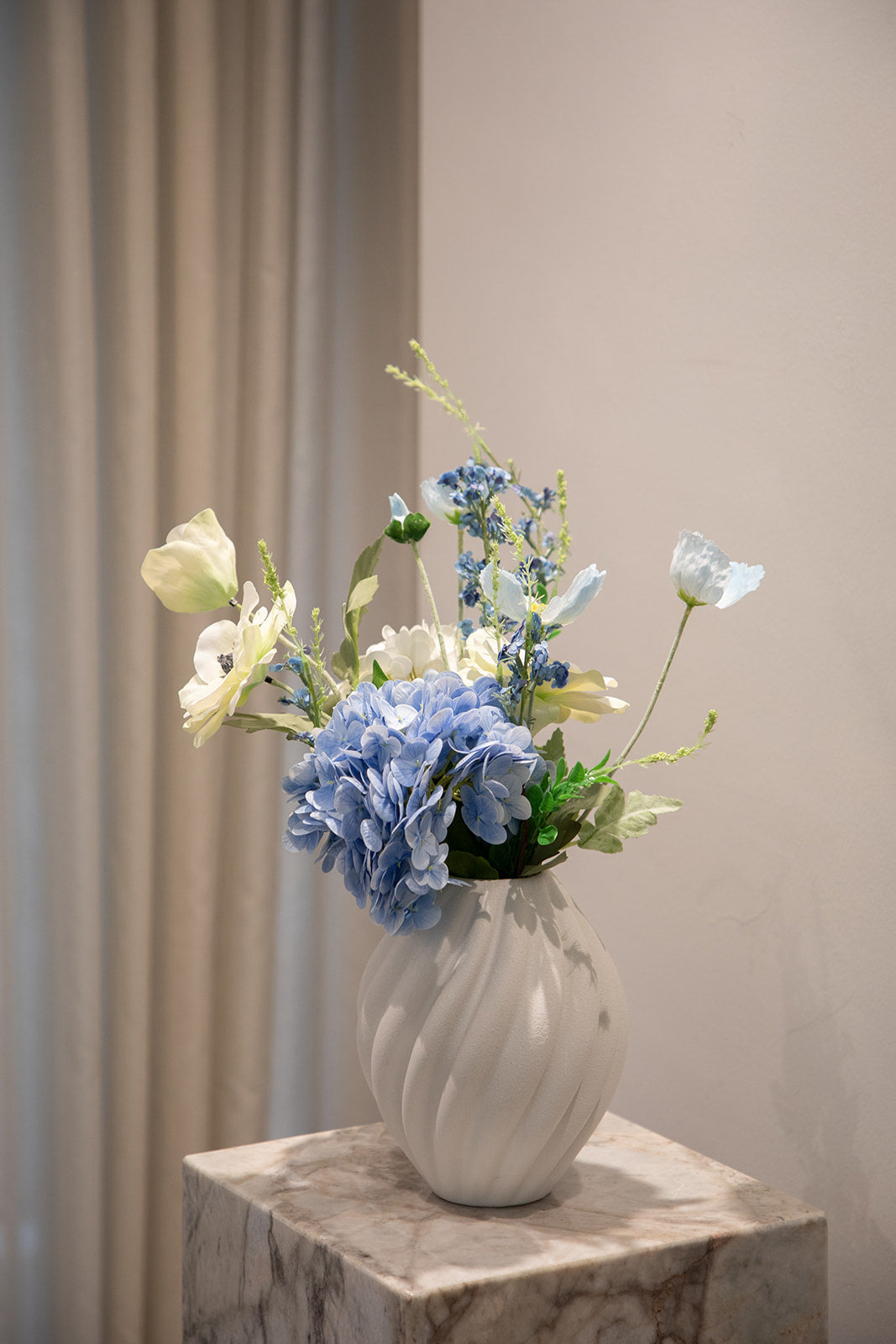 Buket af kunstige blomster i hvide og blå nuancer. Placeret i en hvid keramikvase, der står på en marmor pedestal