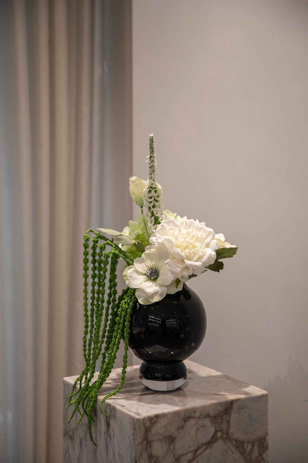 Buket af kunstige blomster i grønne og hvide nuancer. Placeret i en blå glasvase, der står oven på en marmor pedestal.