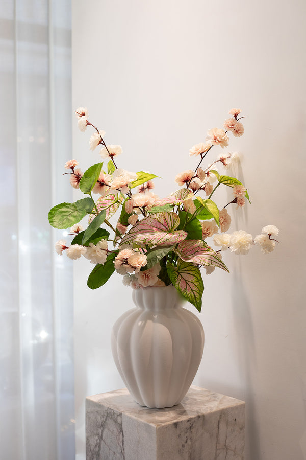 Kunstig buket i sarte rosa, hvide og grønne nuancer. Placeret i en hvid keramik vase, der står på en marmor pedestal.