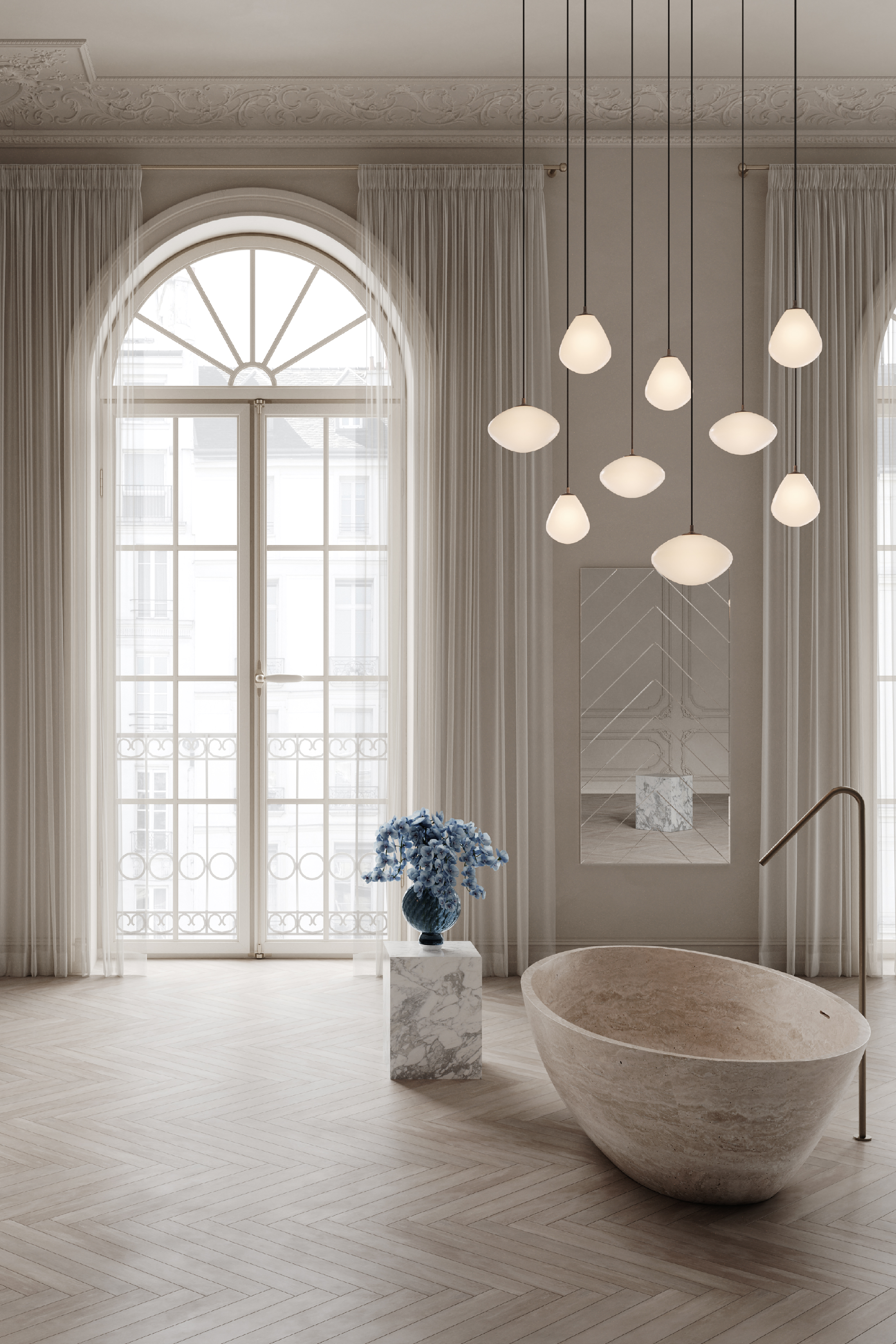 Stort badeværelse med højt til loftet, klassiske stukdetaljer og en fransk altandør med hvide gardiner. Rummet har et fritstående ovalt badekar i natursten, en gulvmonteret messingarmatur og sildebensmønstret trægulv. Over badekarret hænger flere moderne pendellamper med hvide glasskærme. Til venstre står en blå blomsterbuket i en vase på en piedestal af marmor, og bagved ses et vægspejl.