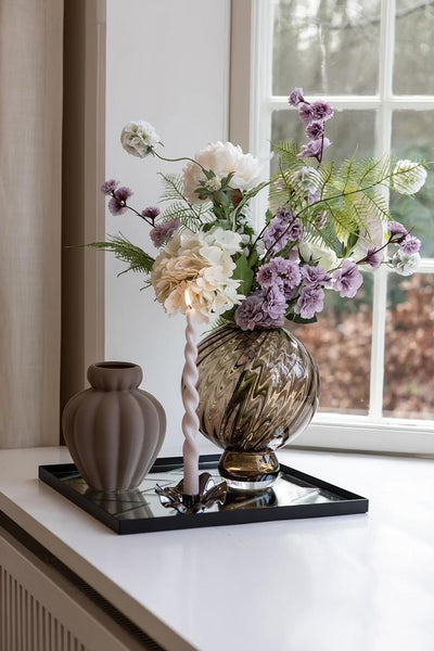 naturtro kunstige blomster i en brun vase fra specktrum placeret i vindueskarm med spejl bakke med en lysestage.