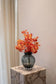 En mørk glasvase med riller, fyldt med en intens buket af røde og orange blomster. Vasen står på en marmorpedestal foran et lyst gardin og en lys væg.