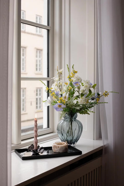 Video af buket af kunstige blomster, der står i en blå glasvase i en vindueskarm. Ved siden af står en bakke med en skål og en lysestage med et snoede lys.